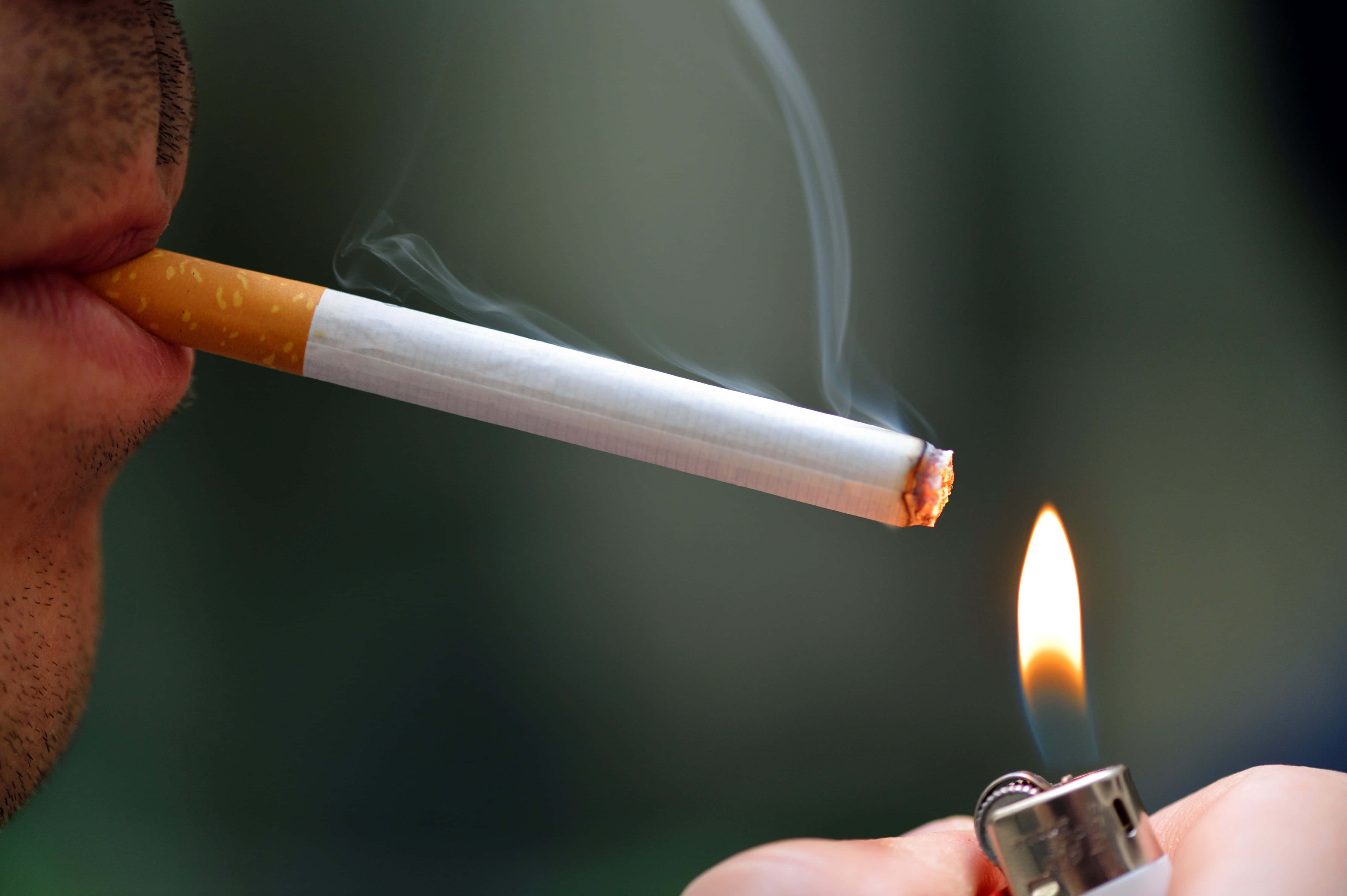 抽根煙，暫時放鬆 壁紙 — 一個人用打火機點燃香煙的特寫，煙霧在模糊的綠色背景上裊裊升起。