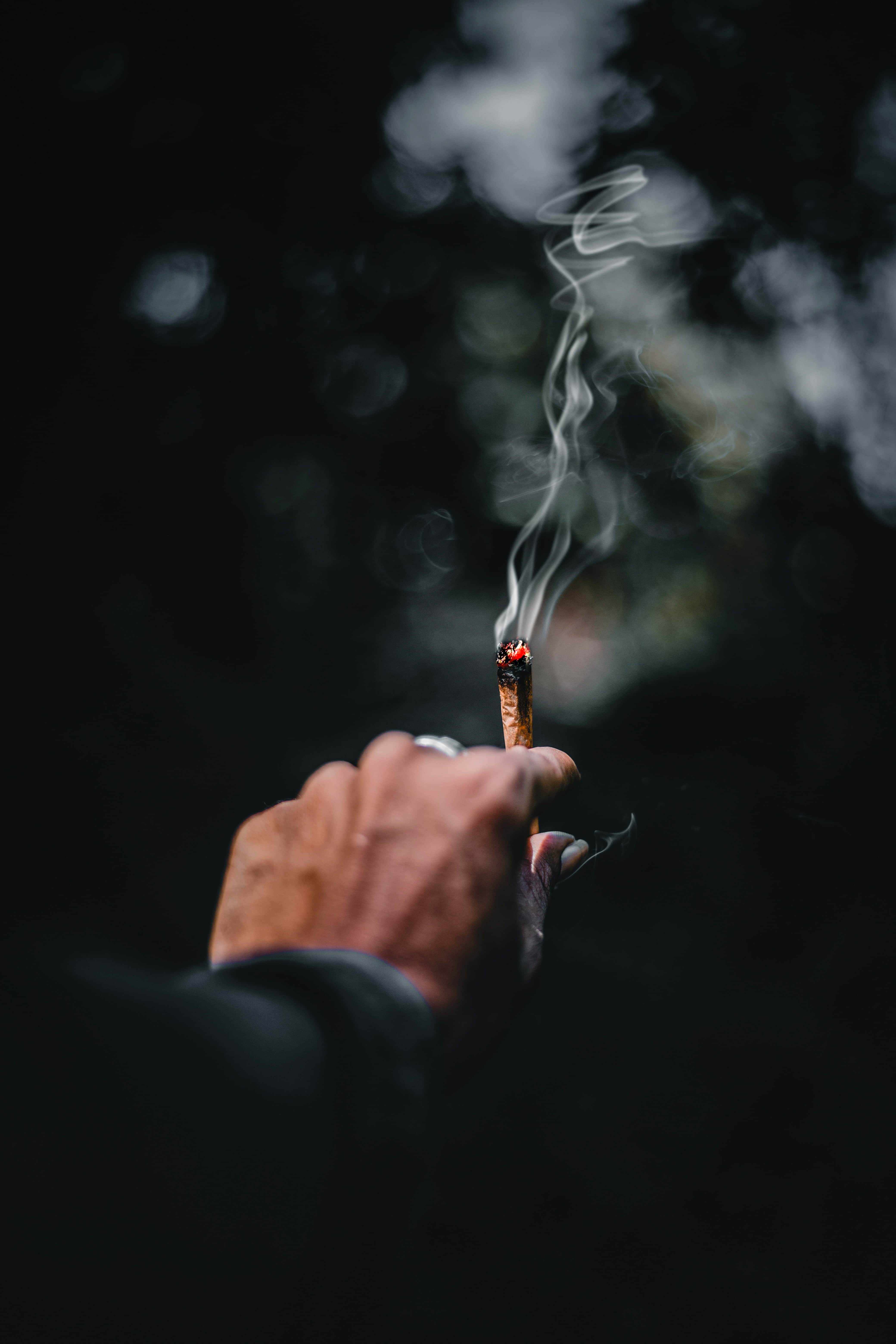 抽煙草的手 壁紙 — 一隻手拿著點燃的香煙，背景模糊黑暗，煙霧向上繚繞。