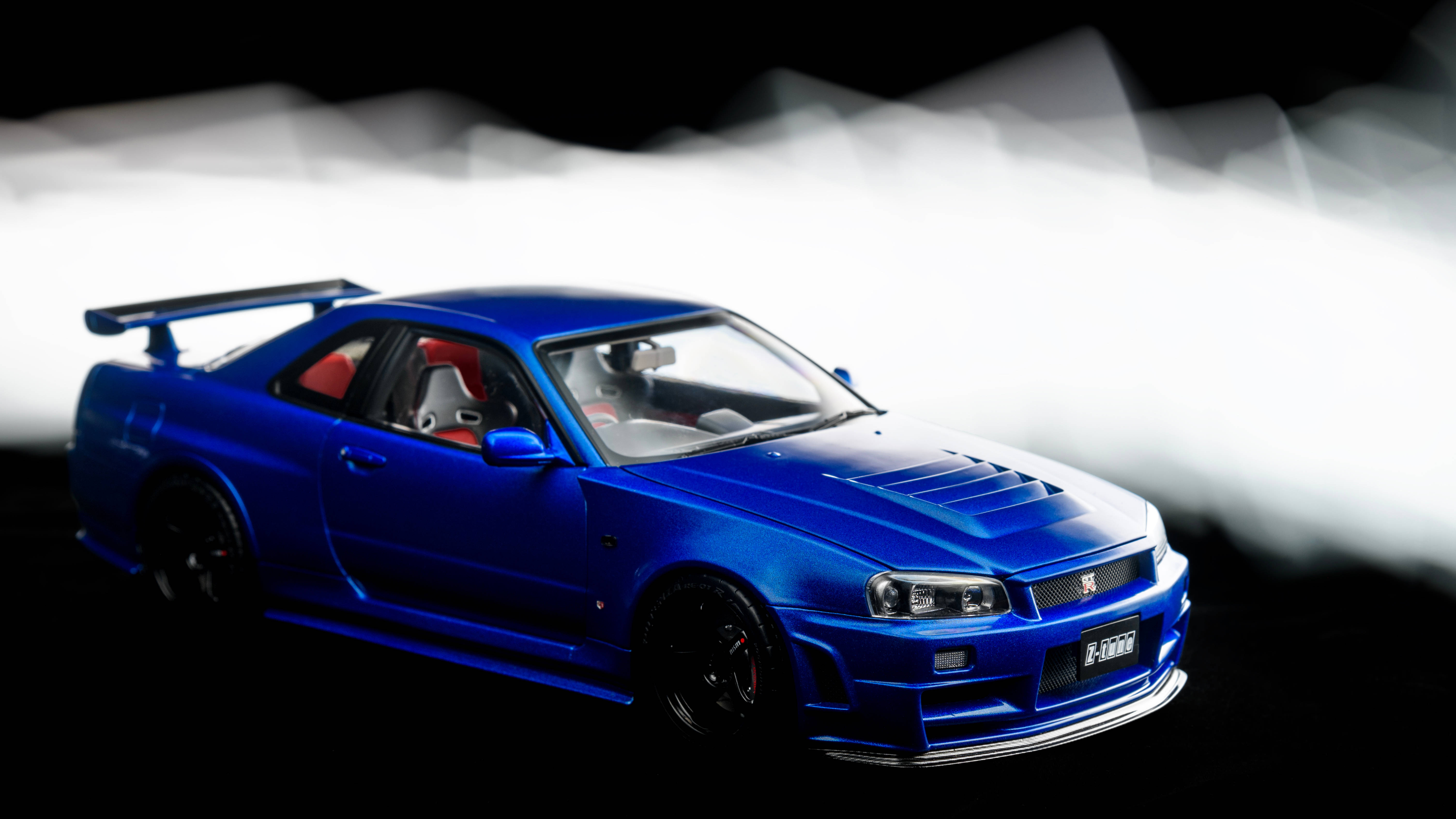 抽象光影 Nissan Skyline GTR R34 壁紙 — 一輛光澤藍色的日產Skyline GT-R模型車，配備紅色內飾和黑色車輪，在煙霧繚繞的白色和黑色背景下戲劇性地照明。
