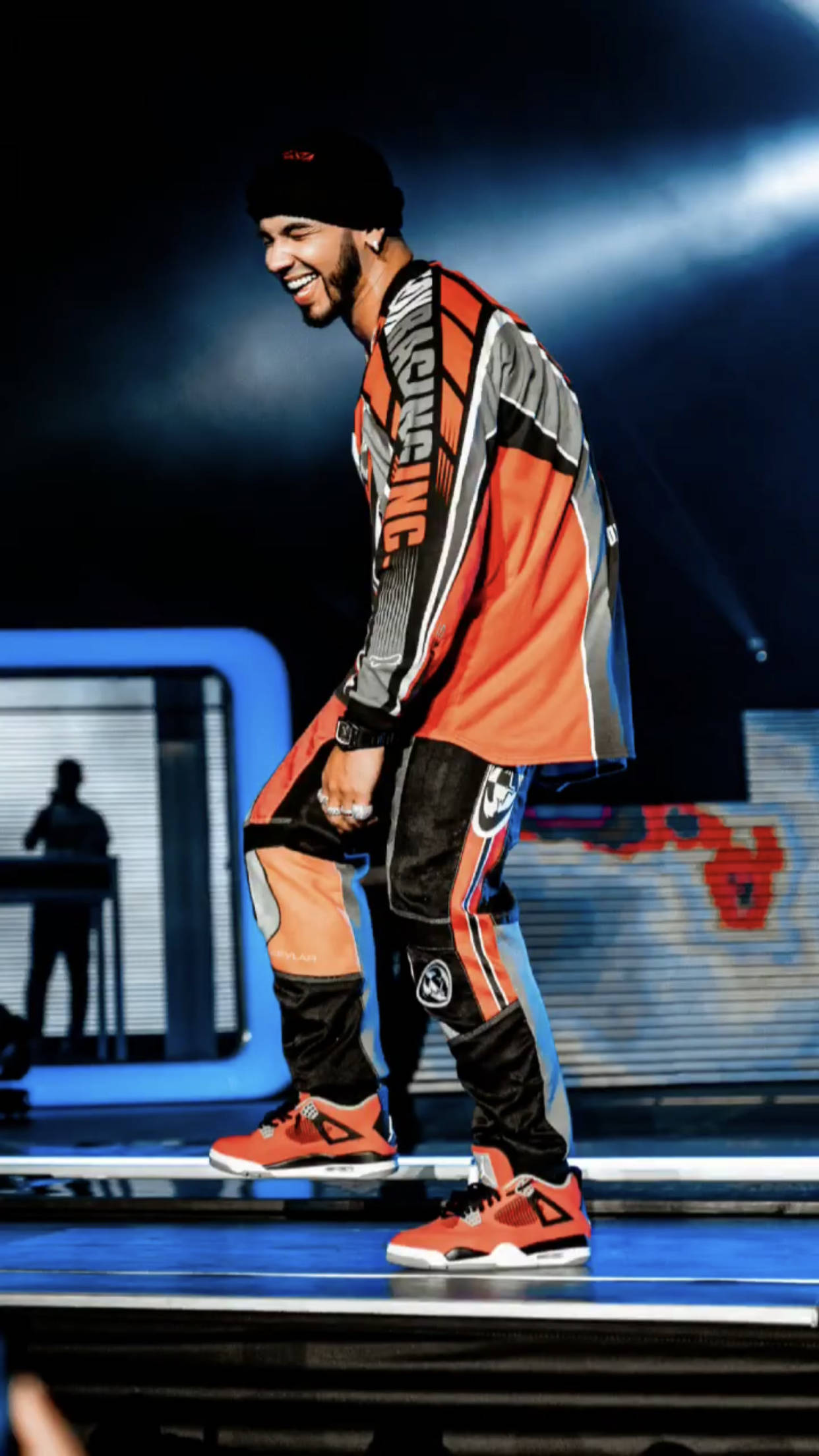 拉丁歌手 Anuel AA 壁紙 — 一位微笑的表演者身穿橙色、灰色和黑色的賽車裝備，在戲劇性的藍色聚光燈下自信地走在明亮的舞台上。
