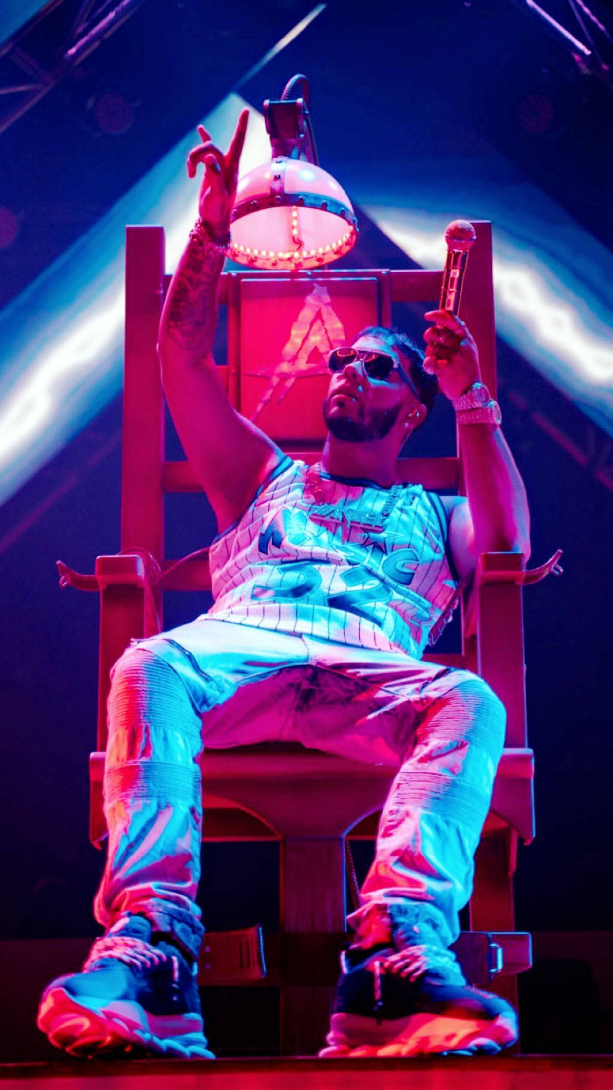 拉丁陷阱雷鬼頓 Anuel AA 壁紙 — 表演者威嚴地坐在王座上，在充滿活力的舞台燈光下，一手做出和平手勢，另一手拿著麥克風。