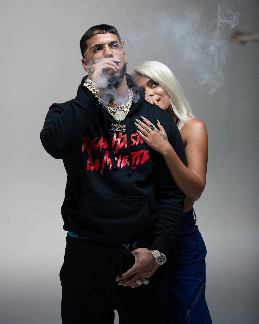 拉丁饒舌歌手 Anuel AA 與 Karol G 壁紙 — 一個穿著黑色連帽衫的男人呼出煙霧，而一個金髮女人在純灰色背景中擁抱他。