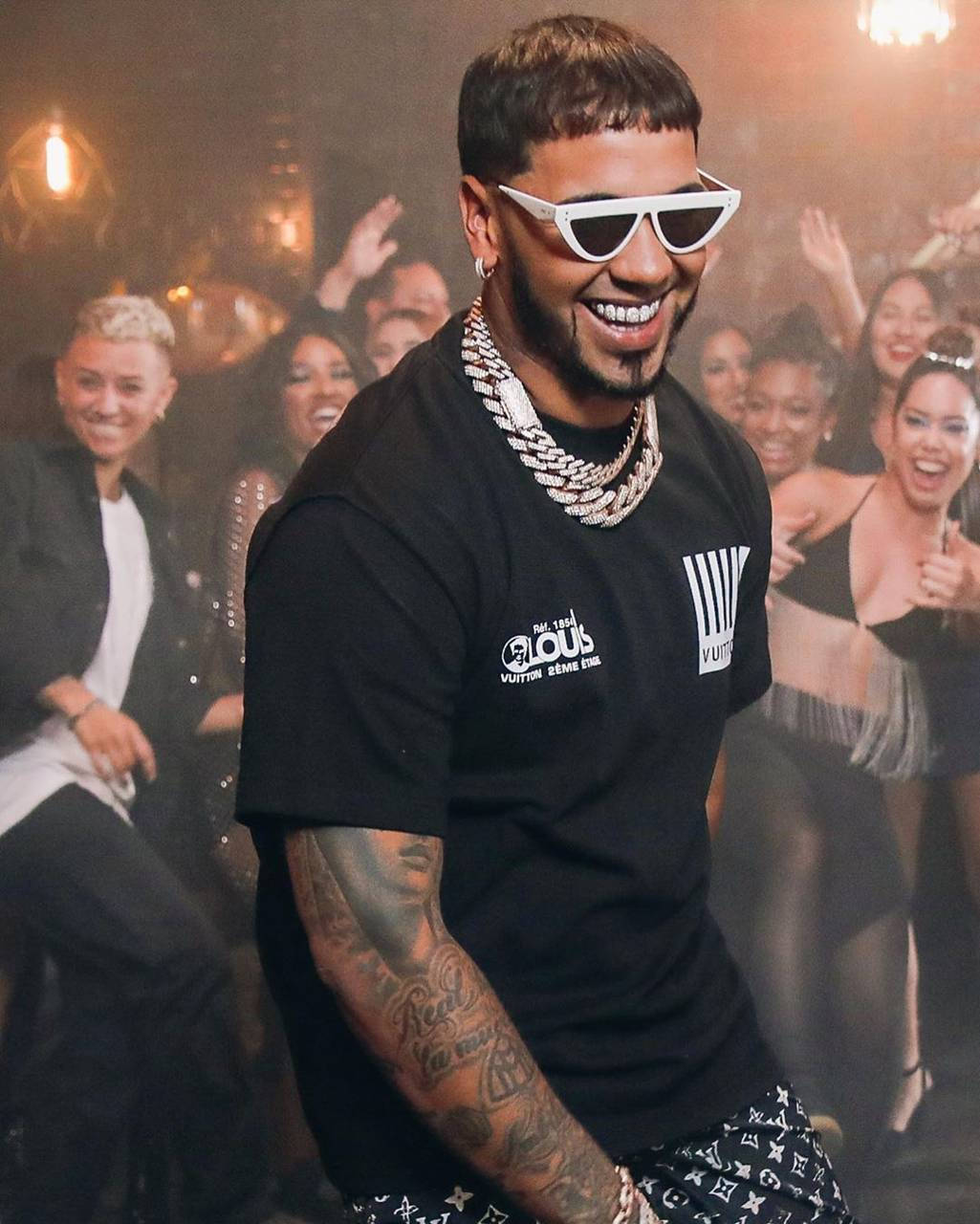 拉丁饒舌歌手 Anuel AA 壁紙 — 一個微笑的男人戴著白色太陽眼鏡和黑色T恤，在熱鬧的派對中自信地擺姿勢，身後有歡呼的人群。