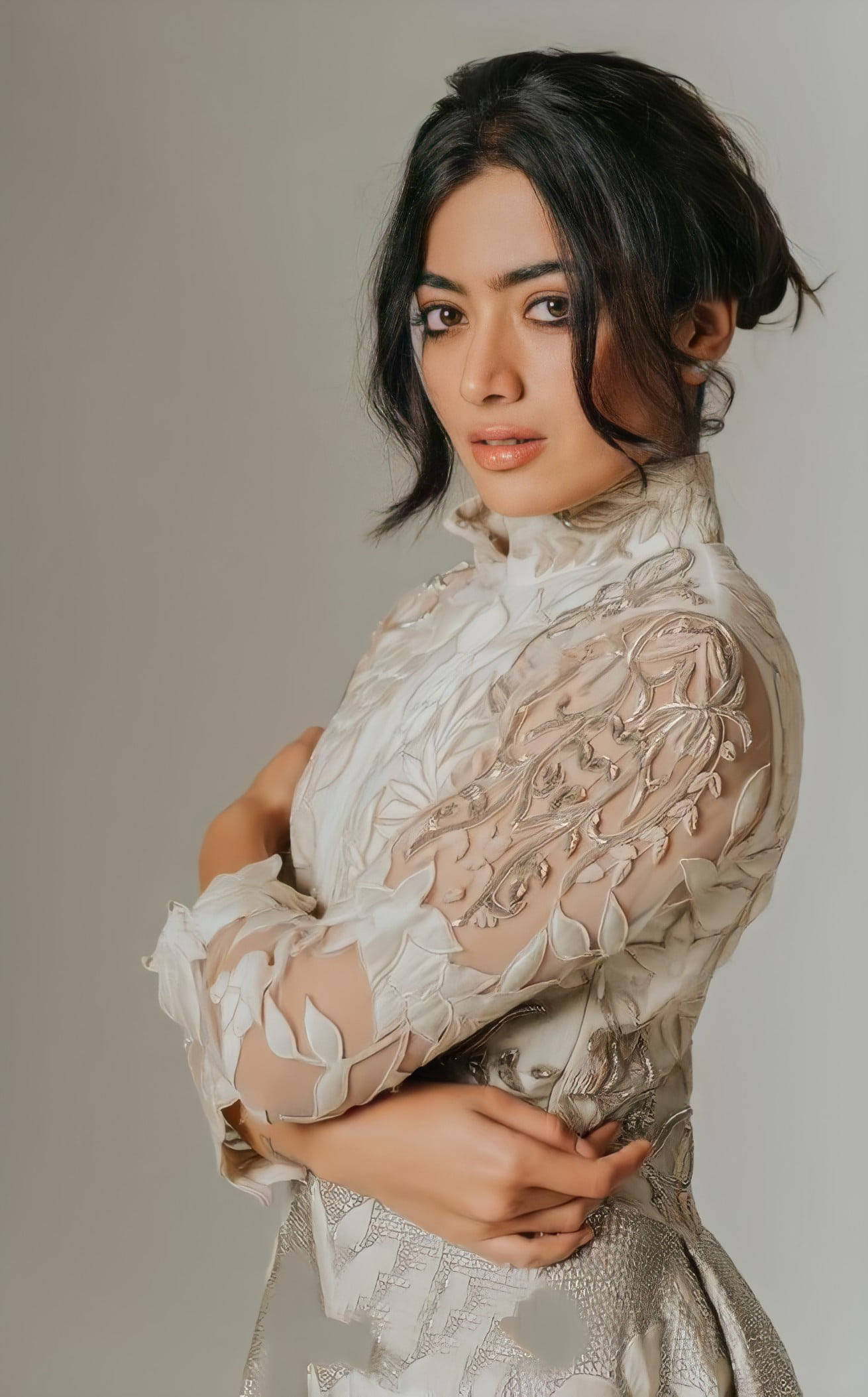 拉什米卡·曼丹納 HD 花邊禮服 壁紙 — 一位優雅的女士穿著華麗的白色蕾絲連衣裙，雙臂交叉，站在中性灰色背景前。