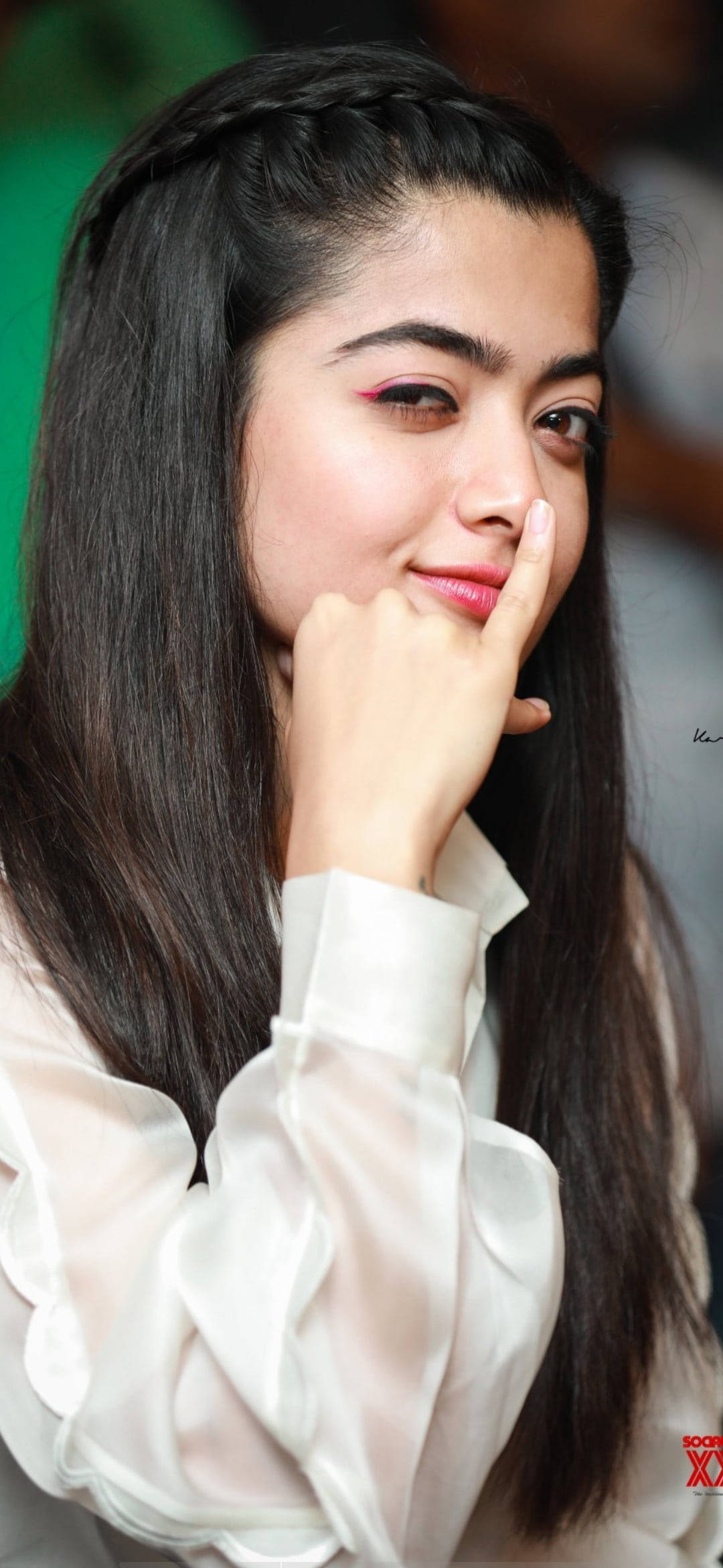 拉什米卡·曼丹納 HD 長黑編髮 壁紙 — 一位年輕女性，長著深色長髮和紅色眼影，將手指放在嘴唇附近，溫柔地微笑。