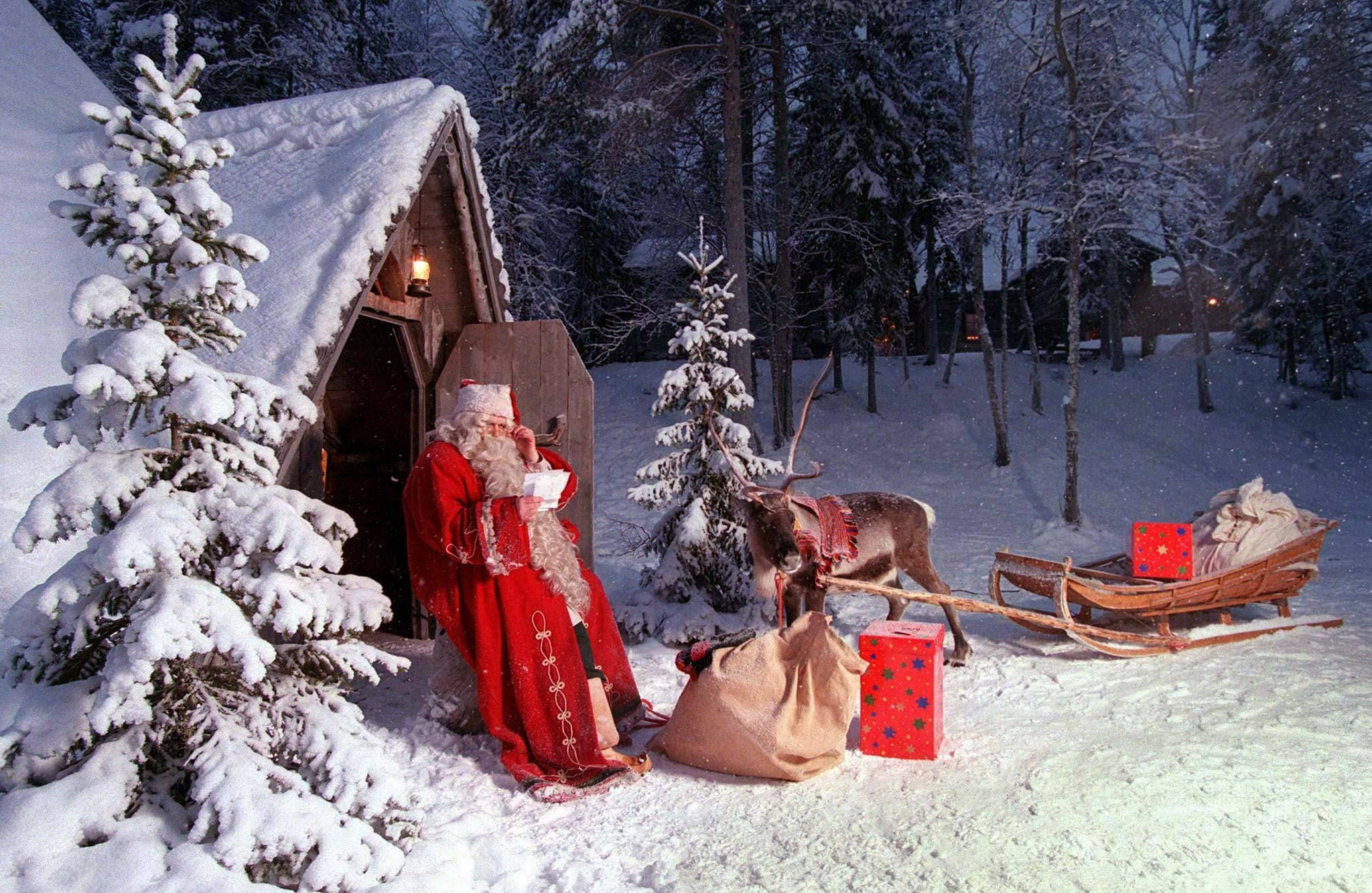 拉普蘭聖誕老人秘境 壁紙 — 身穿紅色長袍的聖誕老人站在雪覆蓋的小屋旁，帶著馴鹿和雪橇，在冬日森林的黃昏時分。