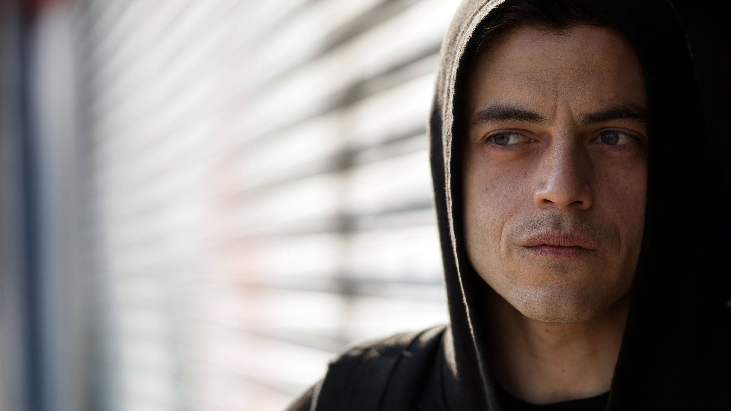 拉米·馬利克在《Mr. Robot》第二季 壁紙 — 一個穿著黑色連帽衫的男人，在模糊的水平百葉窗背景下，帶著嚴肅的表情專注地凝視。
