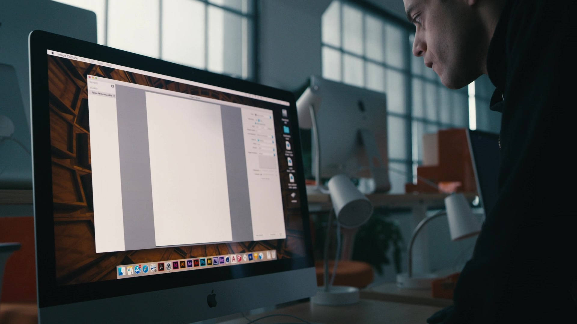 拉米·馬勒克使用iMac 壁紙 — 一個人在燈光昏暗的現代辦公室裡，俯身看著一個大型 iMac 屏幕，屏幕上顯示一個帶有木質背景壁紙的空白文檔。
