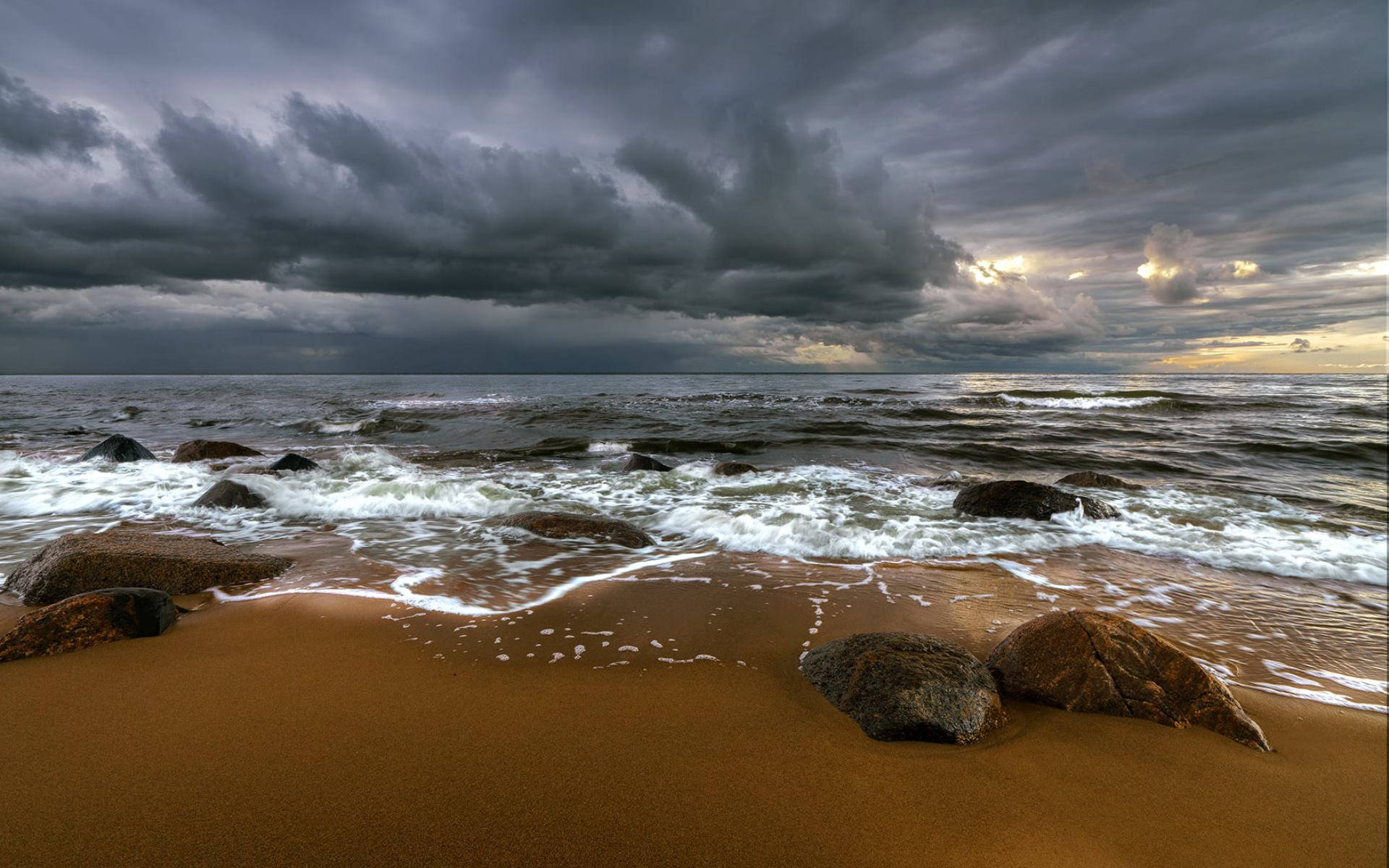 拉脫維亞海景 壁紙 — 暴風雨的海浪拍打沙灘和暗黑岩石，天空烏雲密佈，帶有夕陽餘暉。