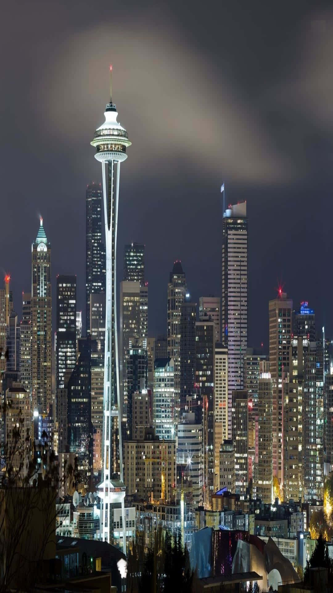 拉長版太空針最佳西雅圖背景 壁紙 — 西雅圖的太空針塔在黑暗多雲的夜空下，在密集的摩天大樓城市景觀上方明亮發光。