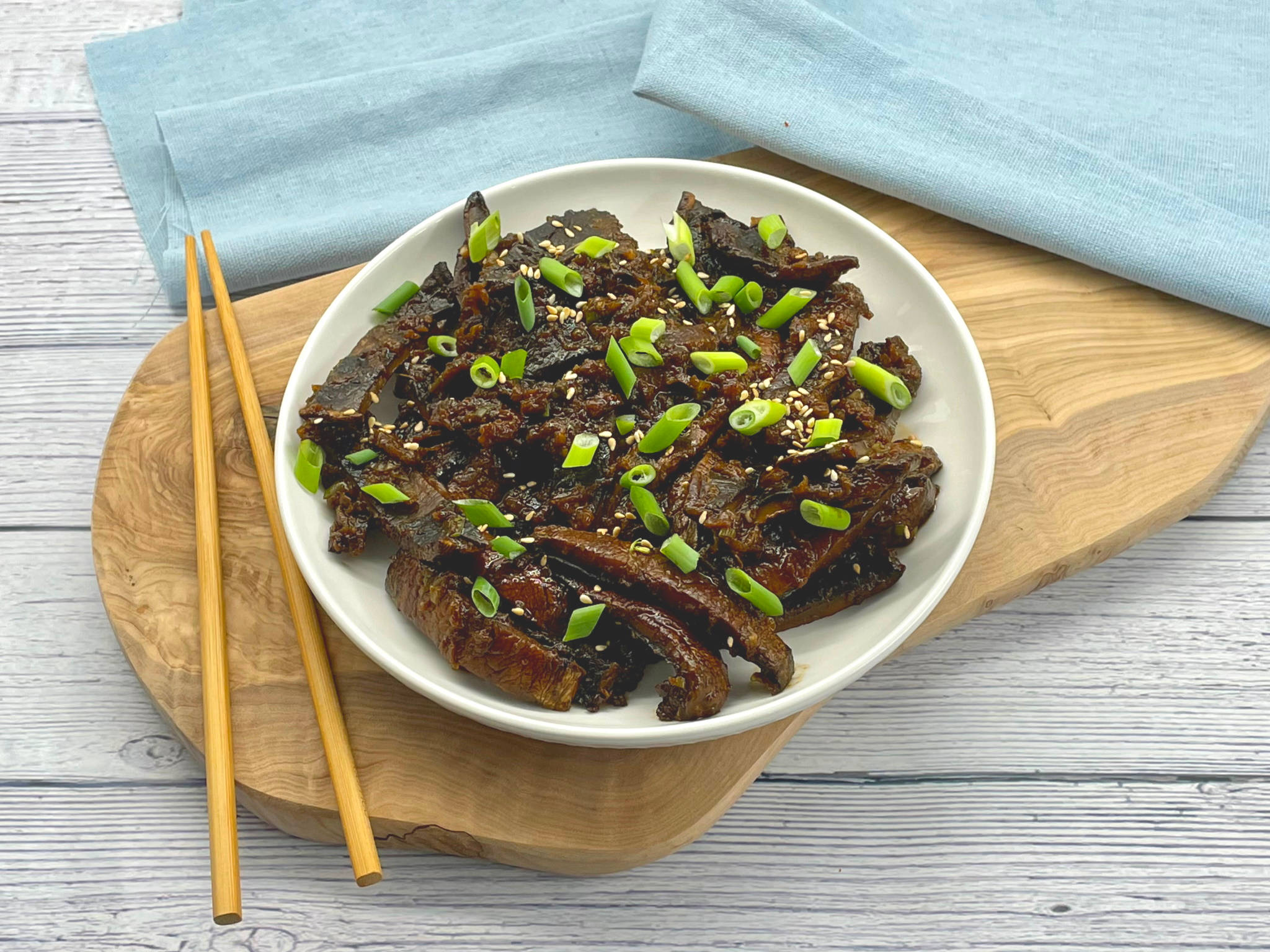 拌醬牛肉配筷子與餐巾 壁紙 — 一個白色碗裡裝著光澤深色釉面鰻魚條，上面撒有蔥花和芝麻，放在木板上，旁邊有筷子，旁邊有一塊藍布。