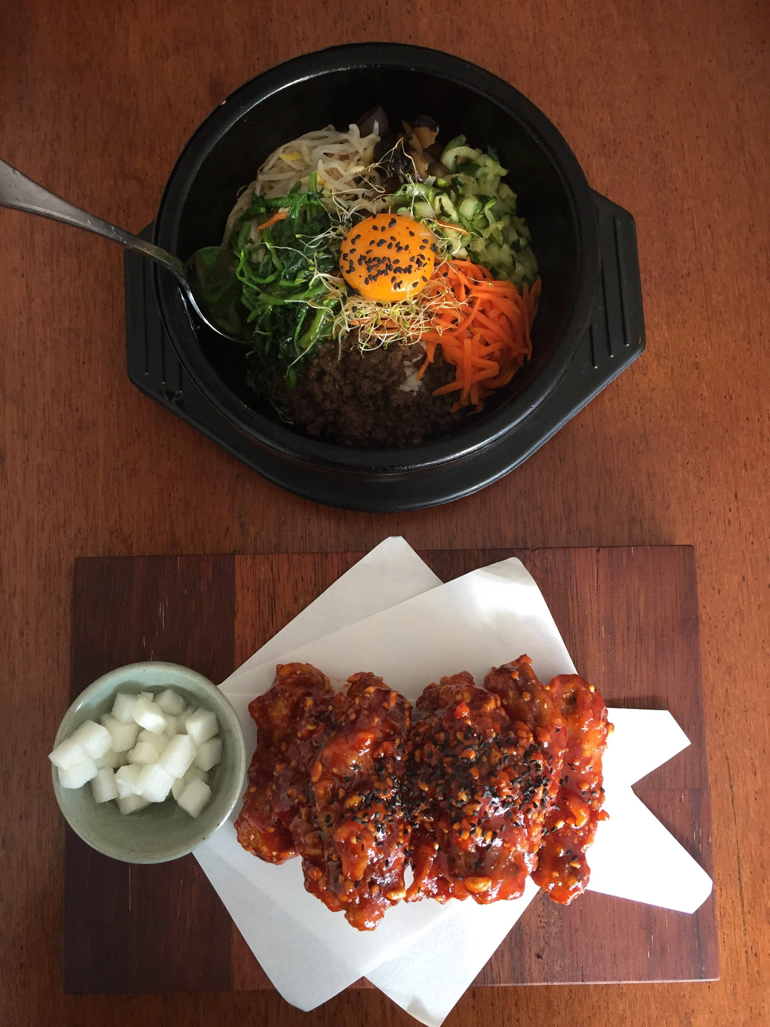 拌飯配照燒雞肉 壁紙 — 俯視角度下，韓國石鍋拌飯配以色彩鮮豔的蔬菜和生蛋黃，旁邊是辣味雞肉和蘿蔔，放在木桌上。