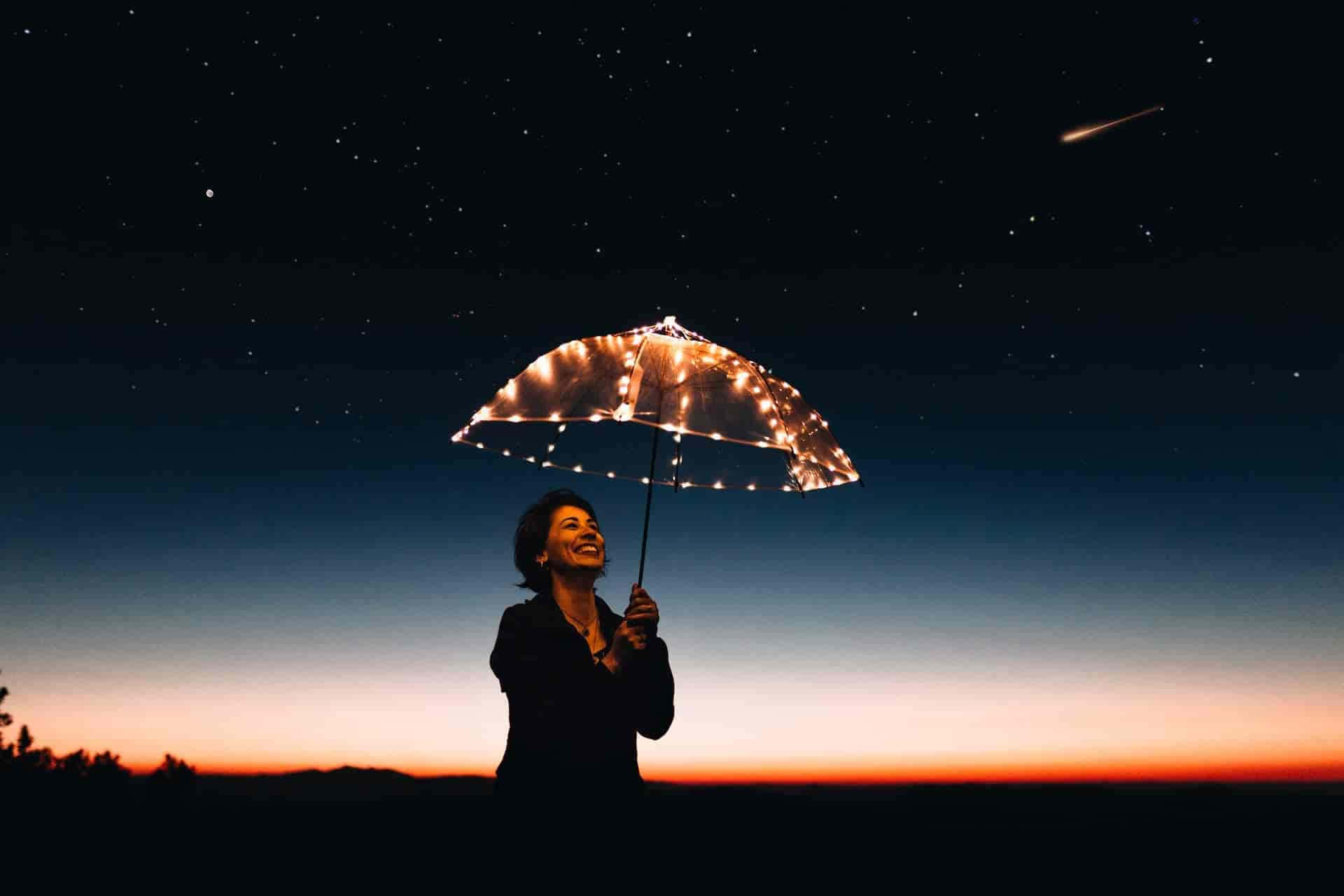 拓展你的想像力 壁紙 — 一位女士在閃爍的燈串傘下微笑，背景是暮色天空，有星星和一顆流星。