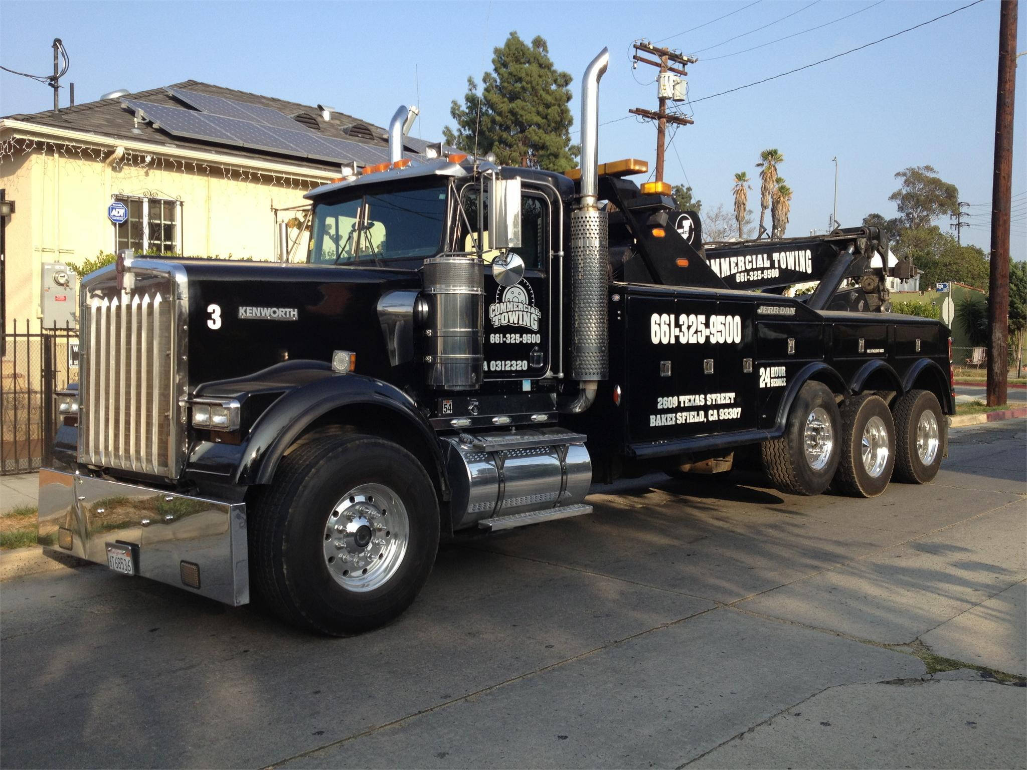 拖曳用凱恩沃斯 W900 壁紙 — 一輛黑色Kenworth拖車，帶有鍍鉻裝飾和可見的拖車設備，停放在陽光明媚的街道上，旁邊是一棟裝有太陽能電池板的房子。