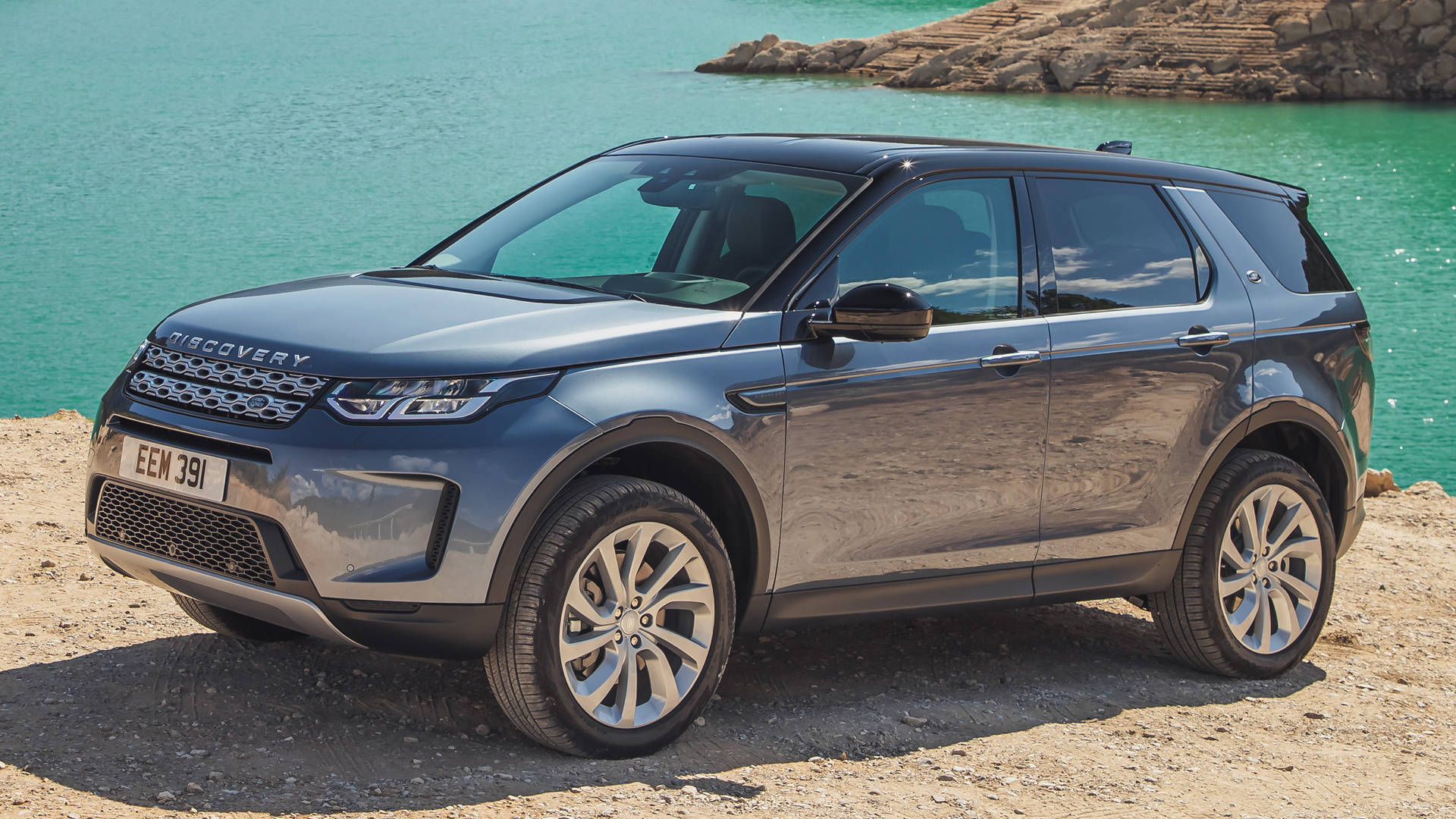 拜倫藍色發現運動版路虎 iPhone 主題壁紙 壁紙 — 一架時尚的灰色 Land Rover Discovery 停泊在陽光普照的岩石海岸旁，旁邊是碧綠色的海水。