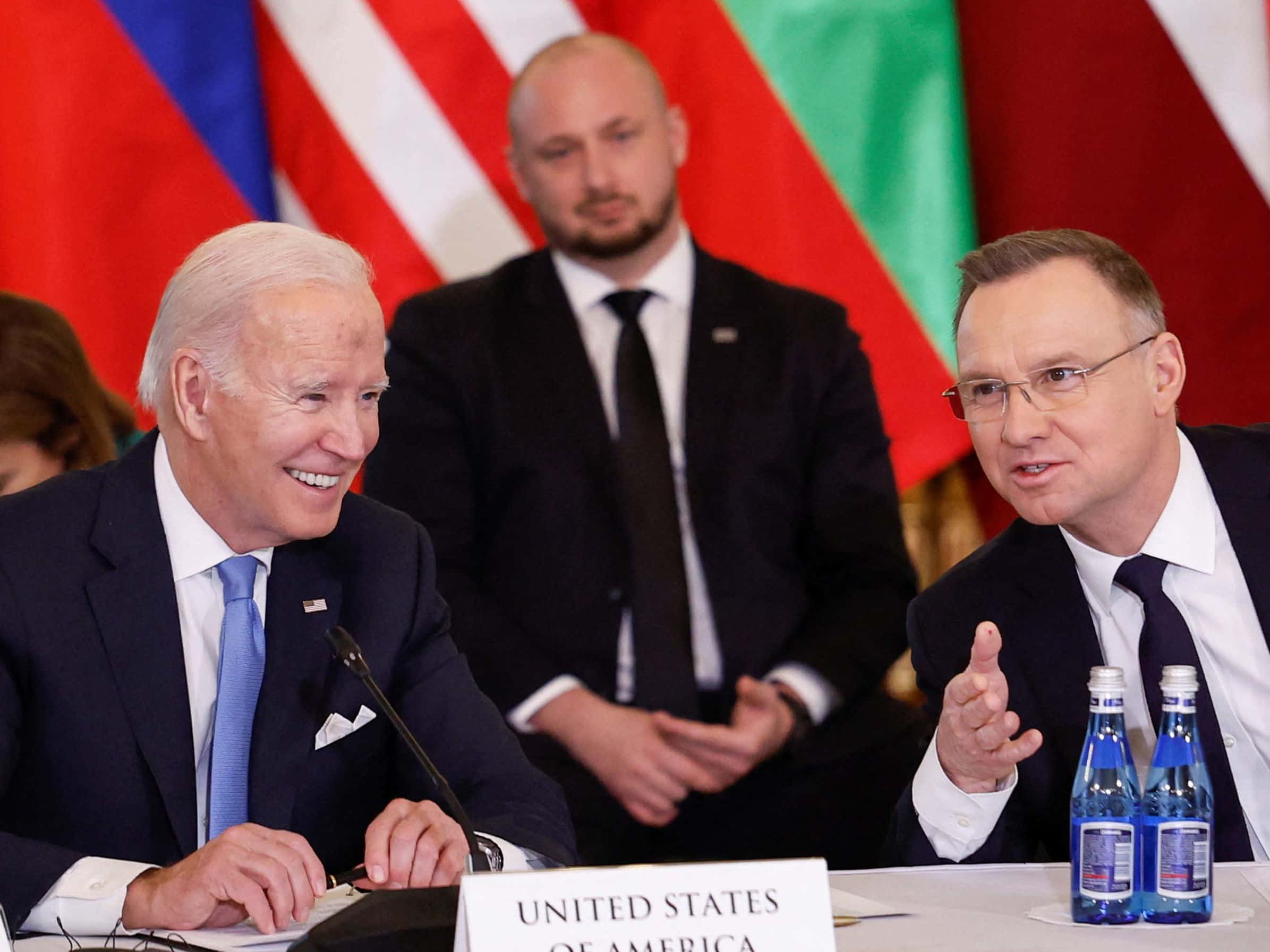 Poland 桌布 壁紙 — 拜登總統微笑著坐在外交會議桌前，桌上放著名牌和樽裝水，另一名男子在正式會議期間做手勢。