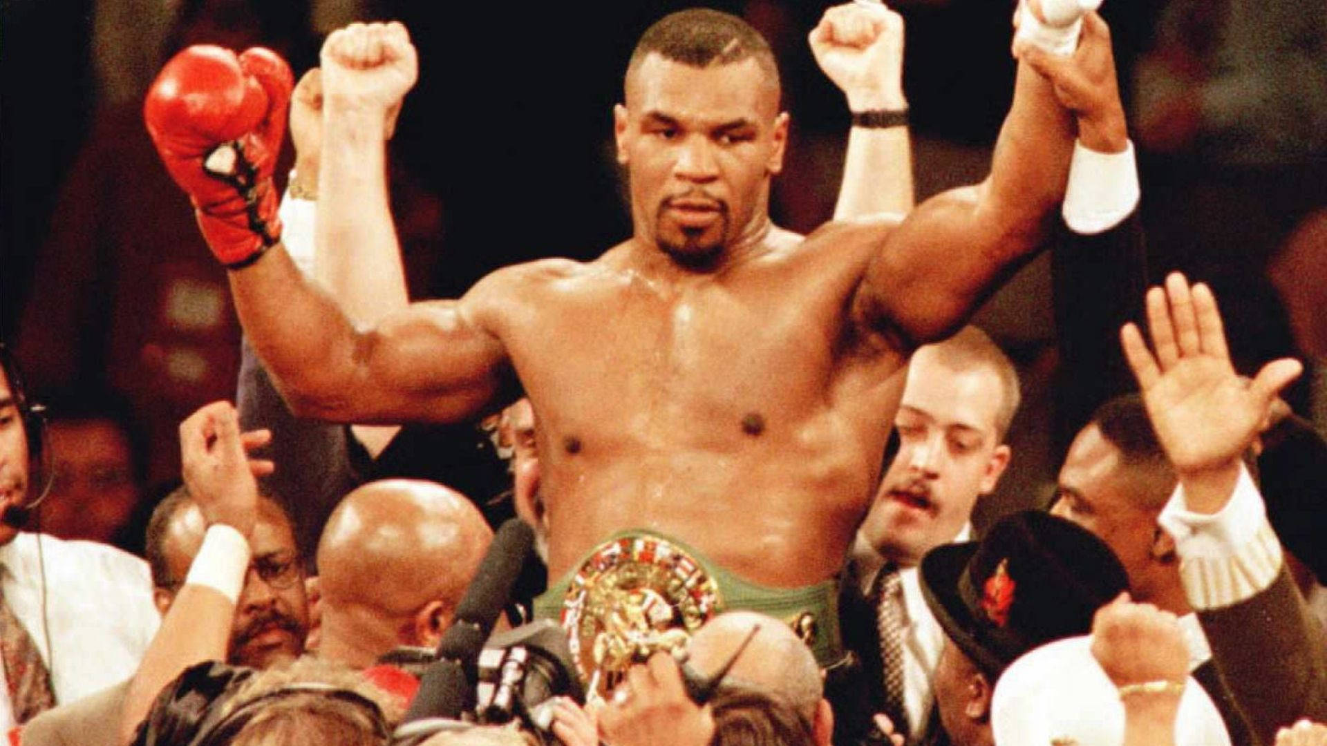 拳王邁克·泰森 壁紙 — Mike Tyson 在擂台上勝利地高舉拳頭，戴著紅色手套和冠軍腰帶，被慶祝的人群包圍。