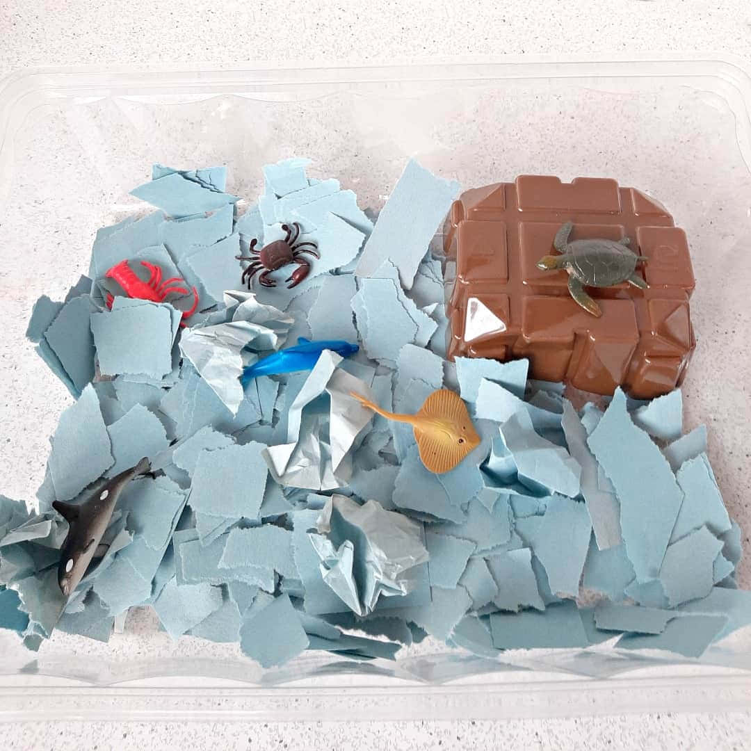 拼湊碎片 壁紙 — 一個透明容器裝有藍色碎紙，內有塑膠海洋生物和一個棕色岩石狀結構。