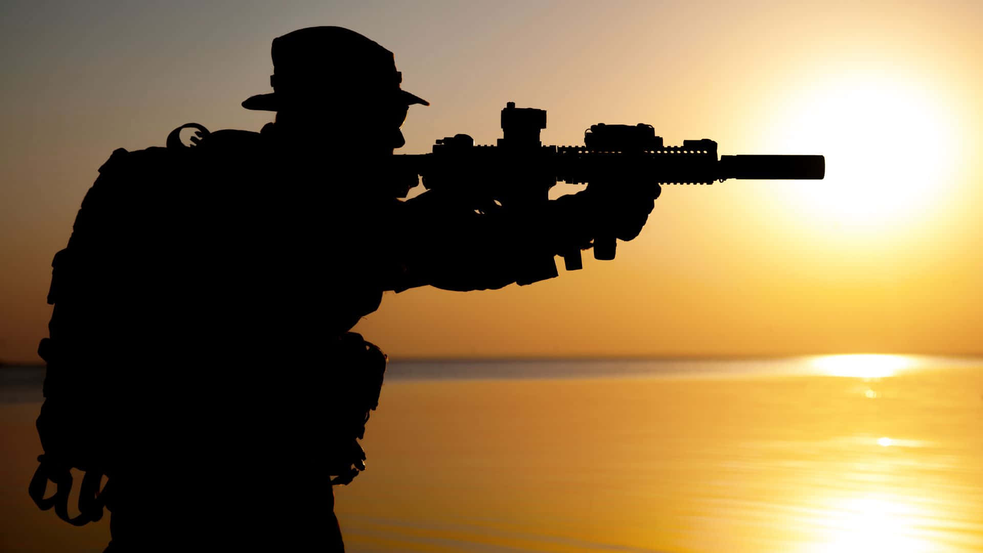 持突擊步槍的軍事作戰人員 壁紙 — 士兵剪影在日落時分於平靜水面上瞄準步槍，帶有金色光芒。