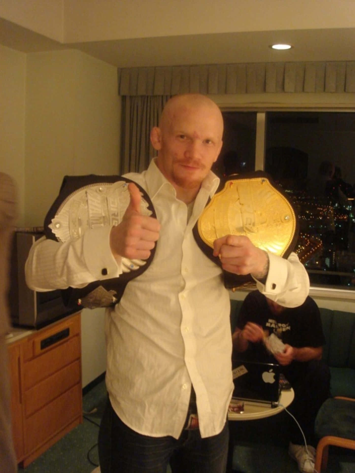 挪威MMA選手約阿希姆·漢森手持兩條冠軍腰帶慶祝勝利 壁紙 — 一位光頭男士身穿白恤衫，在夜晚的酒店房間內自豪地展示兩條冠軍腰帶並豎起大拇指。
