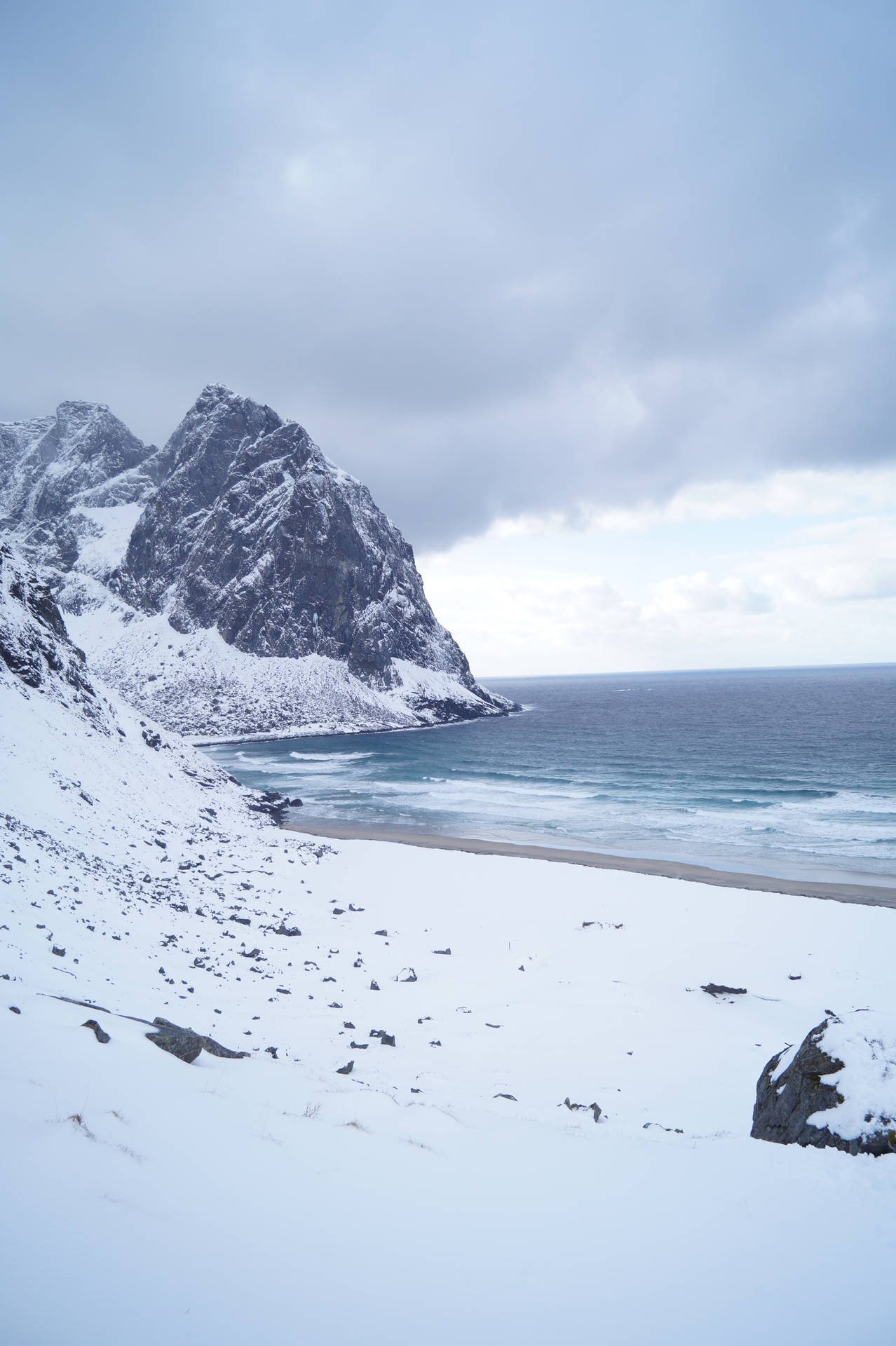 積雪的山崖與沙灘相接，陰雲密布的天空下海浪拍打著海岸。