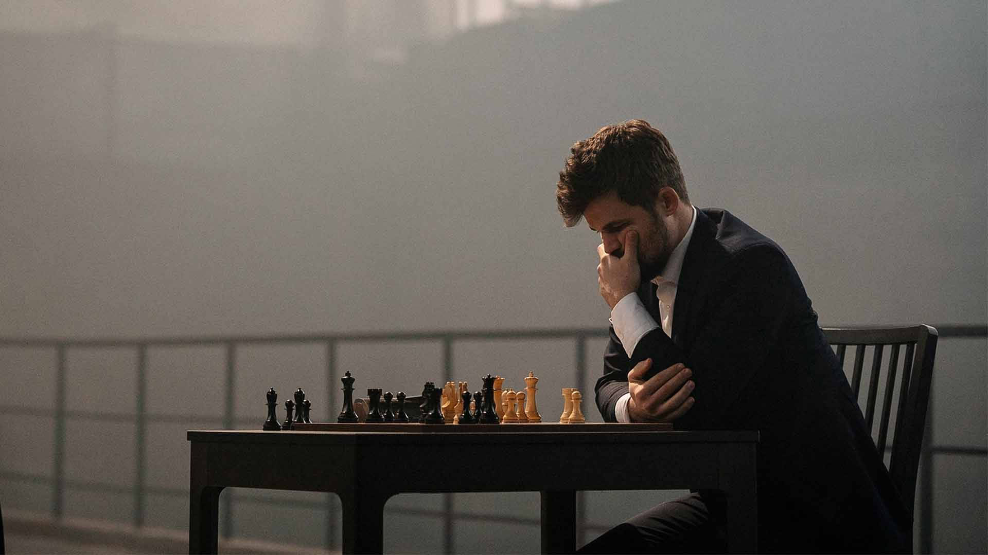 挪威國際象棋大師馬格努斯·卡爾森專注比賽 壁紙 — 一名身穿西裝的男子坐在棋盤桌前，手托下巴陷入沉思，陽光透過他身後朦朧的窗戶灑進來。