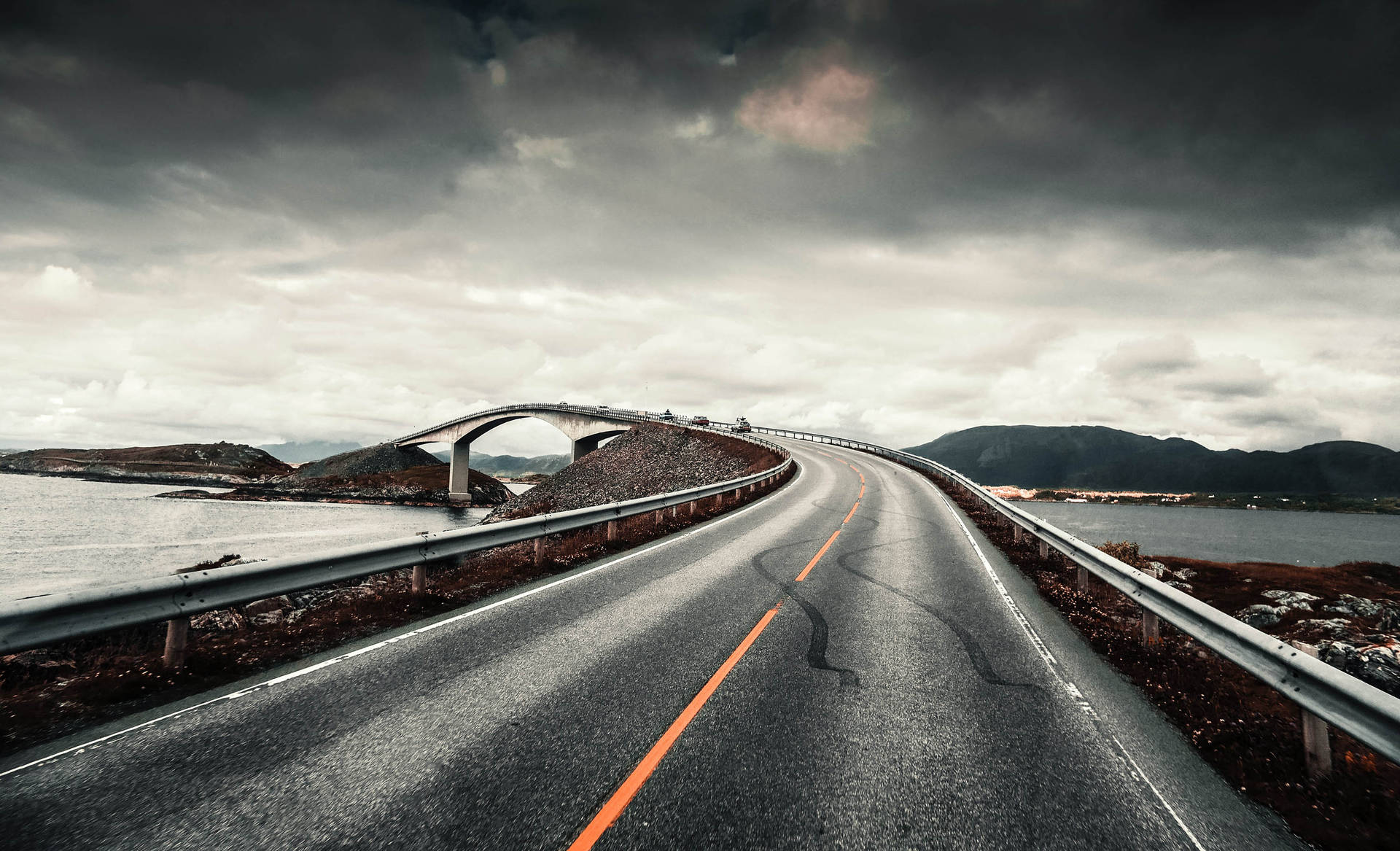 彎曲的海岸道路在風暴天空下通往橋樑，背景有山脈。