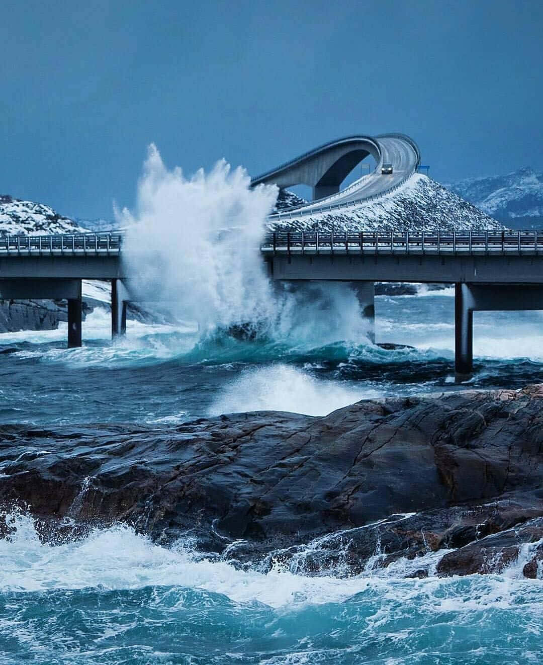 在雪覆蓋的山區海岸景觀中，一道強勁的海浪在拱橋下拍打。