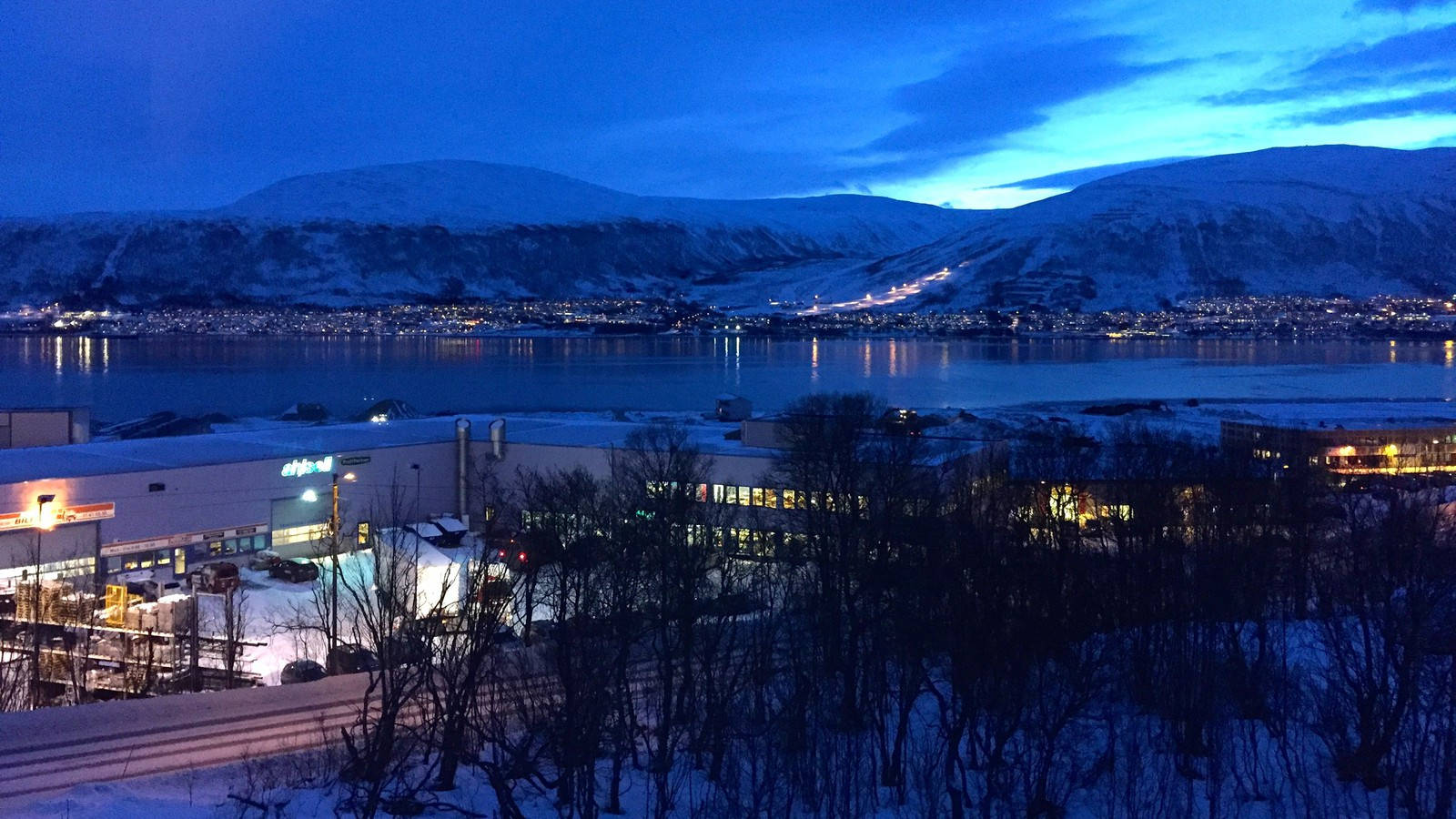 挪威特羅姆瑟極夜景象 壁紙 — 黃昏時分積雪的山城，發光的燈光倒映在平靜的峽灣和前景的禿樹上。