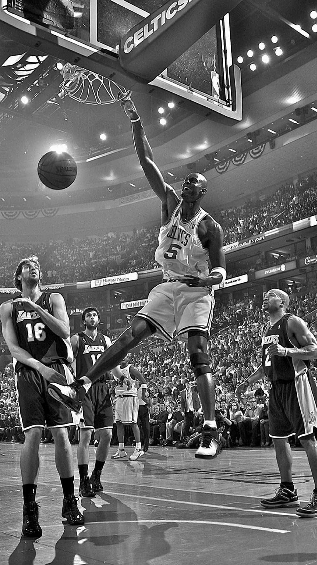 捕捉NBA的精髓！ 壁紙 — 一名身穿塞爾特人球衣的籃球運動員在半空中入樽，對手在擠滿觀眾的場館內觀看。