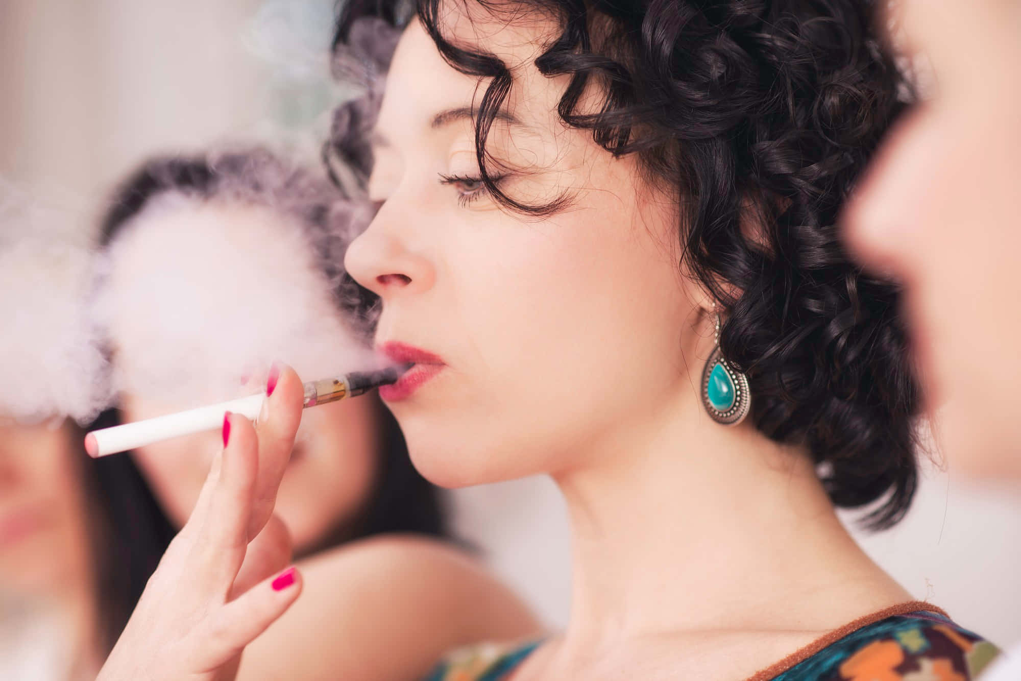 捲髮女性吸電子煙圖 壁紙 — 一位擁有捲曲深色頭髮的女性，戴著綠松石耳環和紅色指甲油，呼出電子煙的蒸氣。