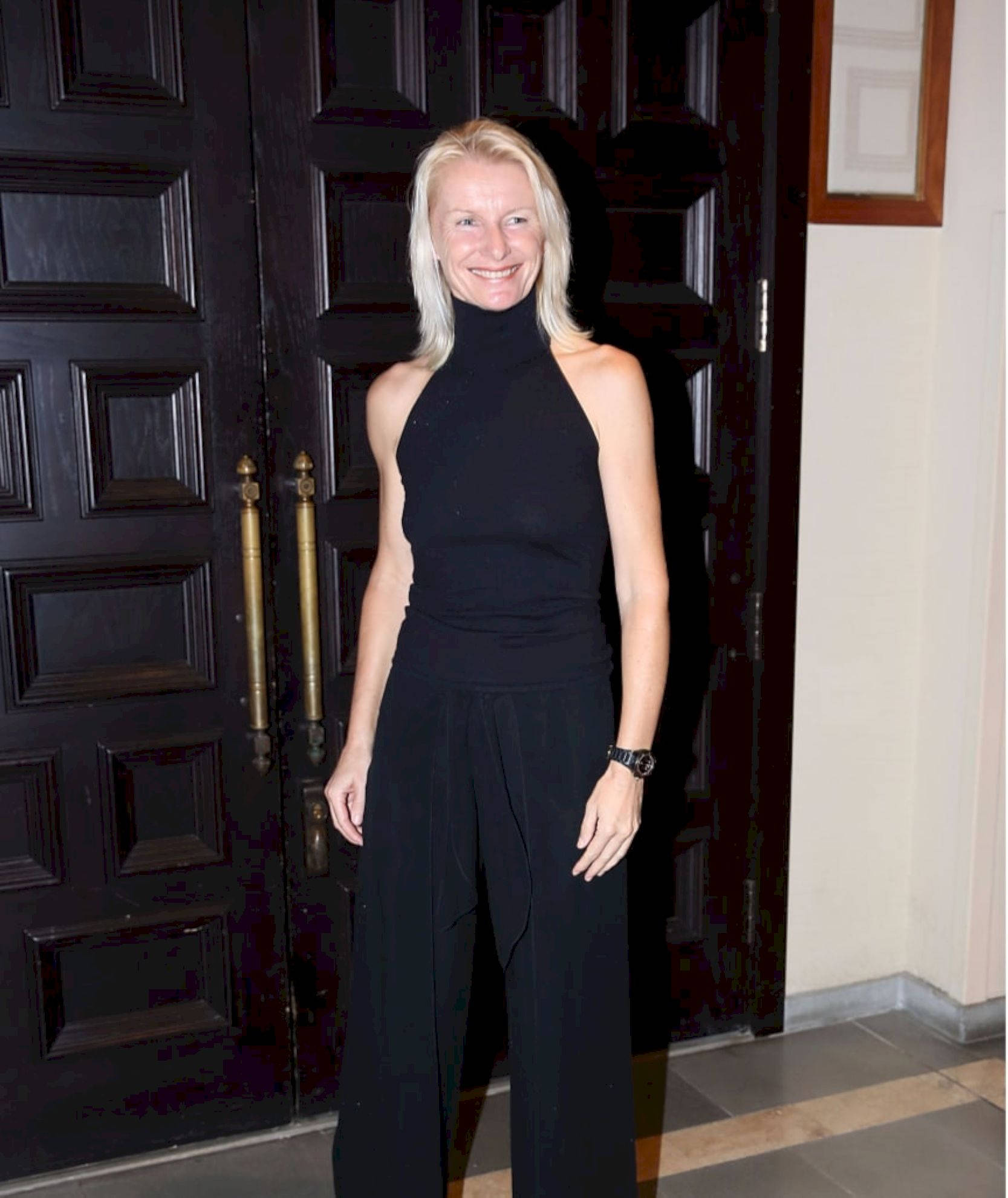 捷克網球傳奇 Jana Novotna 身著優雅黑裙 壁紙 — 一位微笑的金髮女性穿著黑色掛頸上衣和闊腿褲，站在一扇帶有黃銅把手的深色木門前。