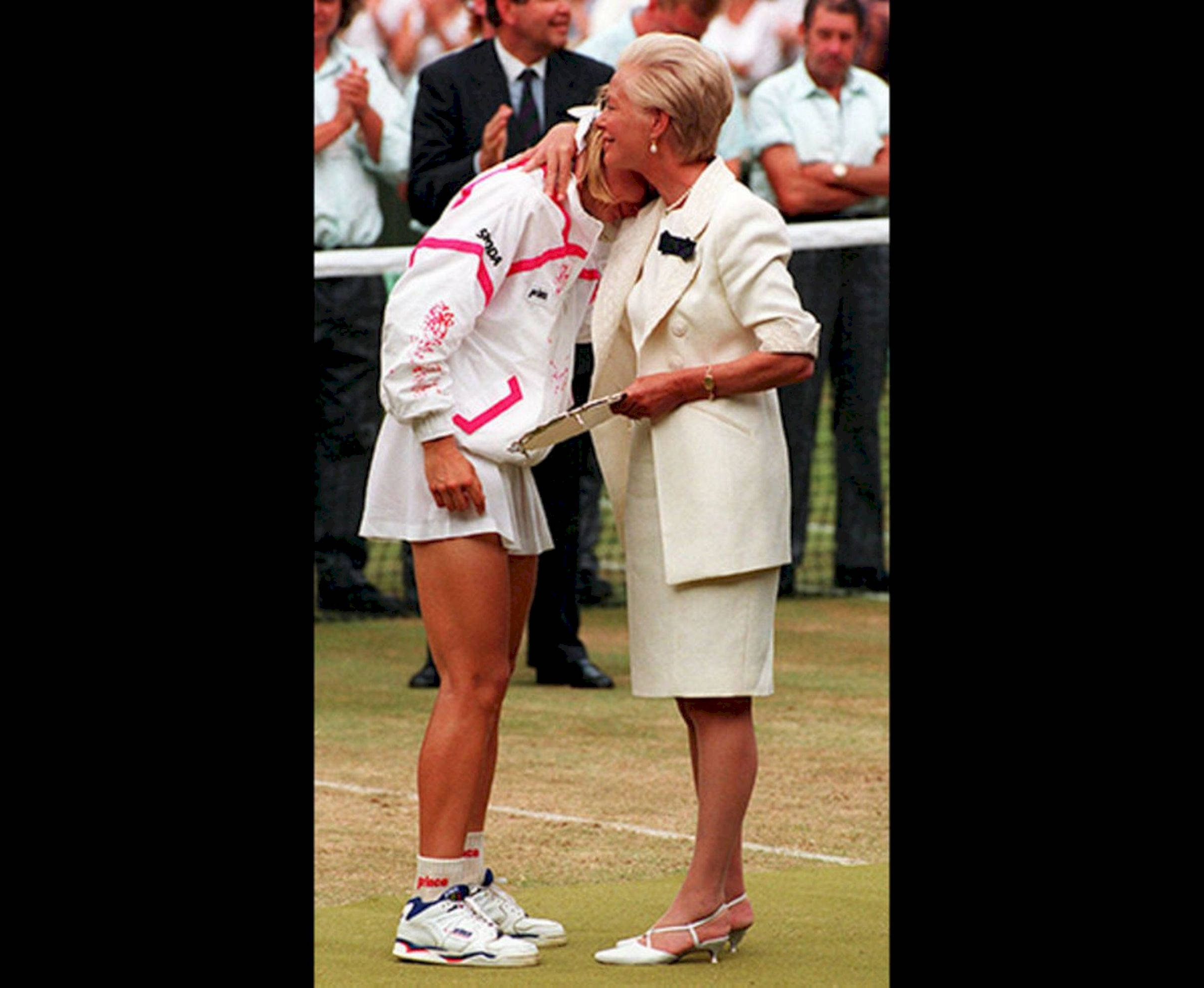 捷克網球名將 Jana Novotna 與肯特公爵夫人 壁紙 — 一名身穿白色和粉紅色運動服的網球運動員在草地場上擁抱一位身穿奶油色西裝的女士，觀眾鼓掌。