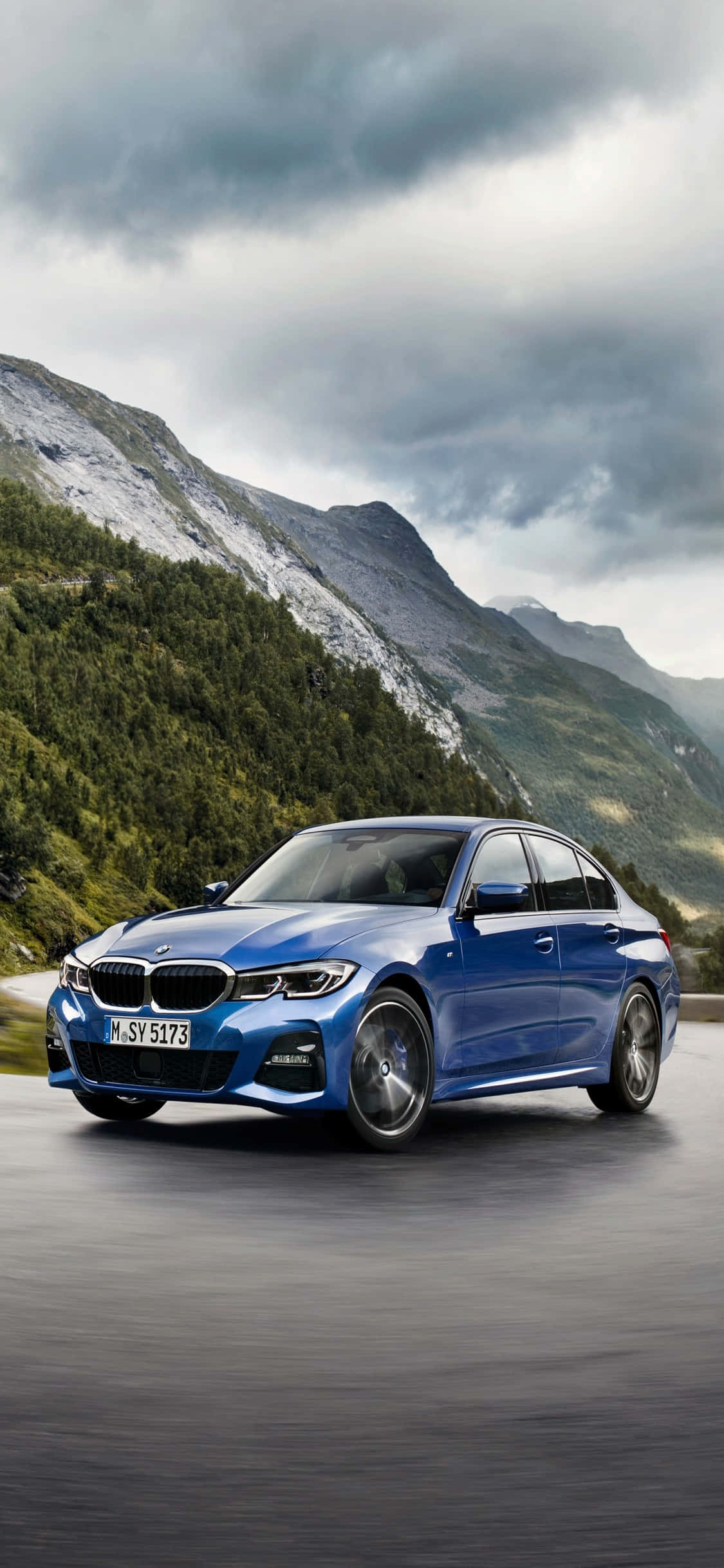 掌中奢華，BMW 的標誌就在 iPhone X 背面 壁紙 — 一輛時尚的藍色BMW轎車在戲劇性的多雲天空下行駛在山路上。