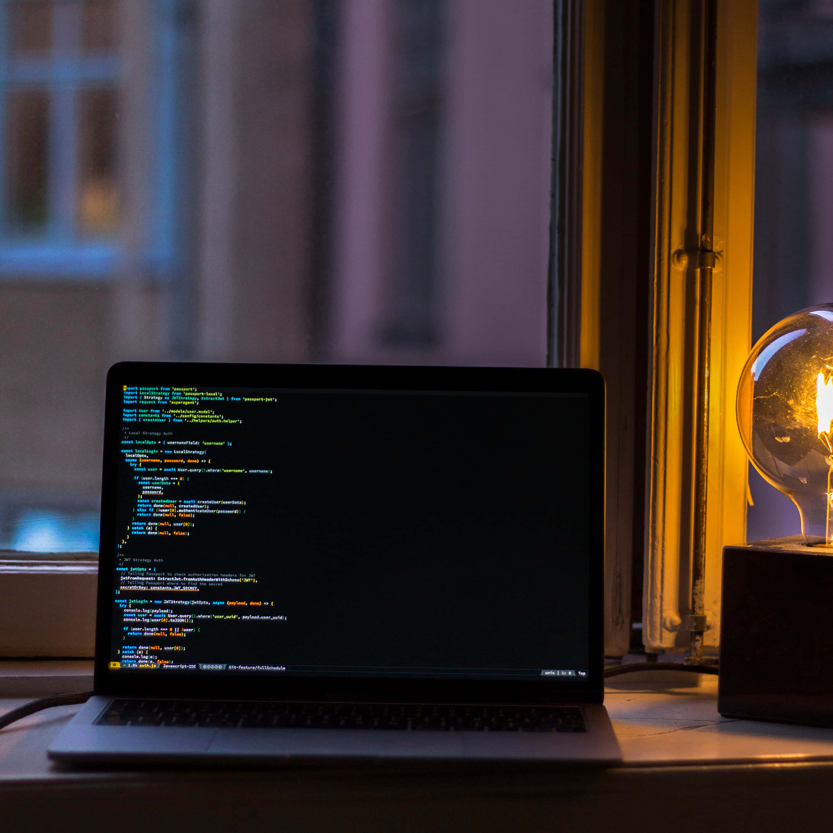 掌握科技的力量 壁紙 — 一部顯示彩色代碼的筆記型電腦放在窗台上，旁邊有一個發光的復古燈泡散發出溫暖的光線。