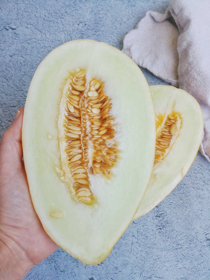 排毒活力黃瓜瓜片 壁紙 — 一隻手拿著半個哈密瓜，露出多汁的淡色果肉和琥珀色的種子，背景是帶紋理的藍色表面。