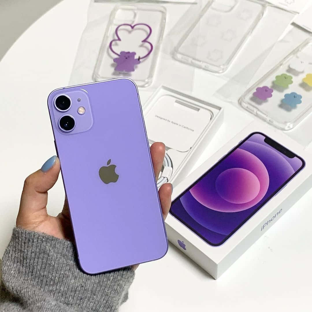探索 iPhone 12 的全新功能 壁紙 — 一隻手拿著啞光紫色 iPhone 12 mini，旁邊是它的盒子和帶有彩色雲朵吊飾的透明保護殼。