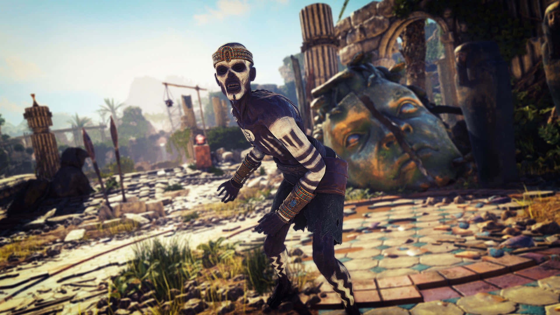 探索 Strange Brigade 的古代遺跡 壁紙 — 一個身穿部落盔甲的骷髏戰士，在陽光明媚的廢墟中，伴隨著巨大的石頭頭顱和破碎的柱子，威風凜凜地站立著。