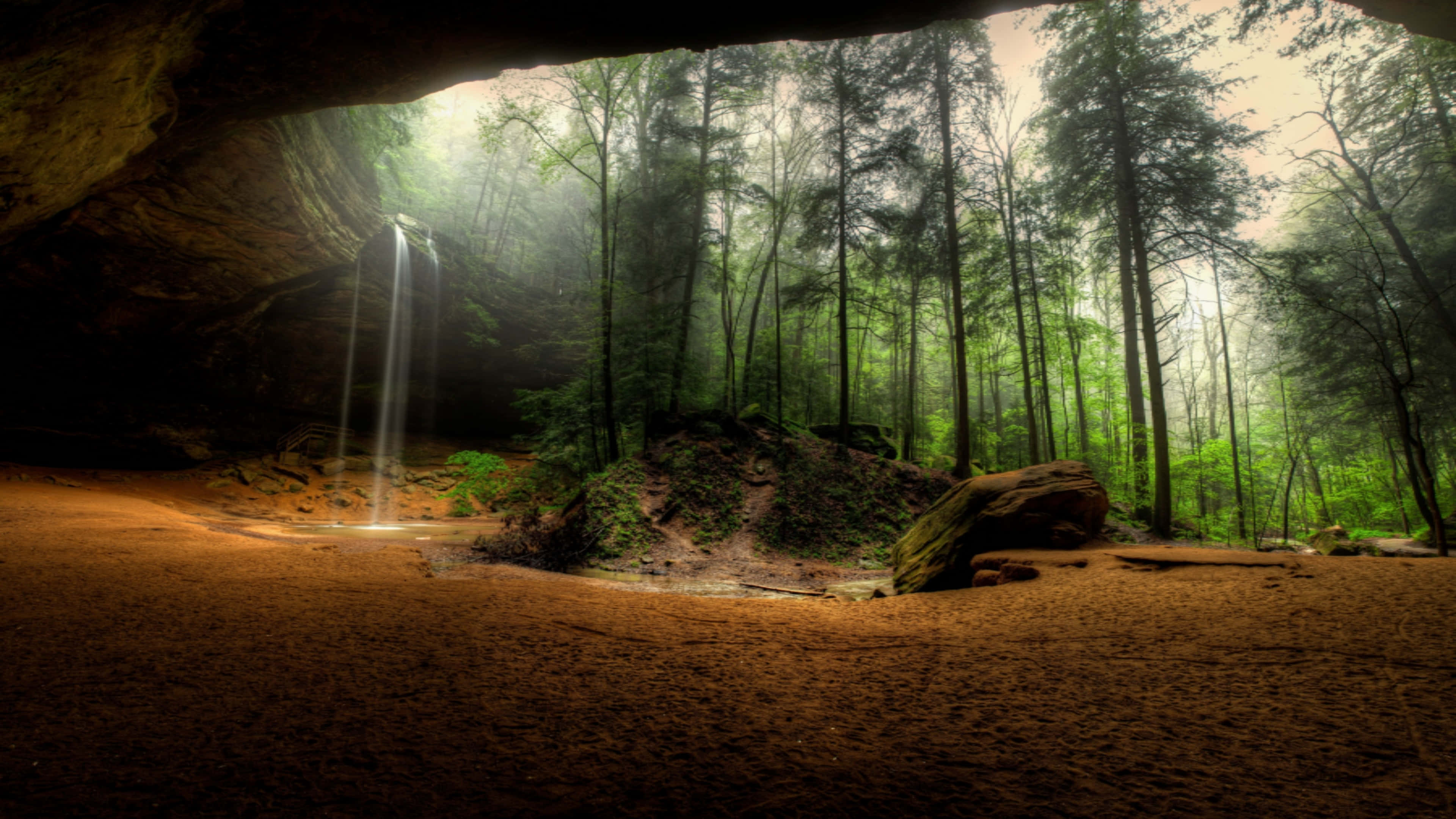 探索 UHD 風景之美 壁紙 — 一個寧靜的森林瀑布從洞穴開口流入一個籠罩在柔和金光中的霧氣森林。