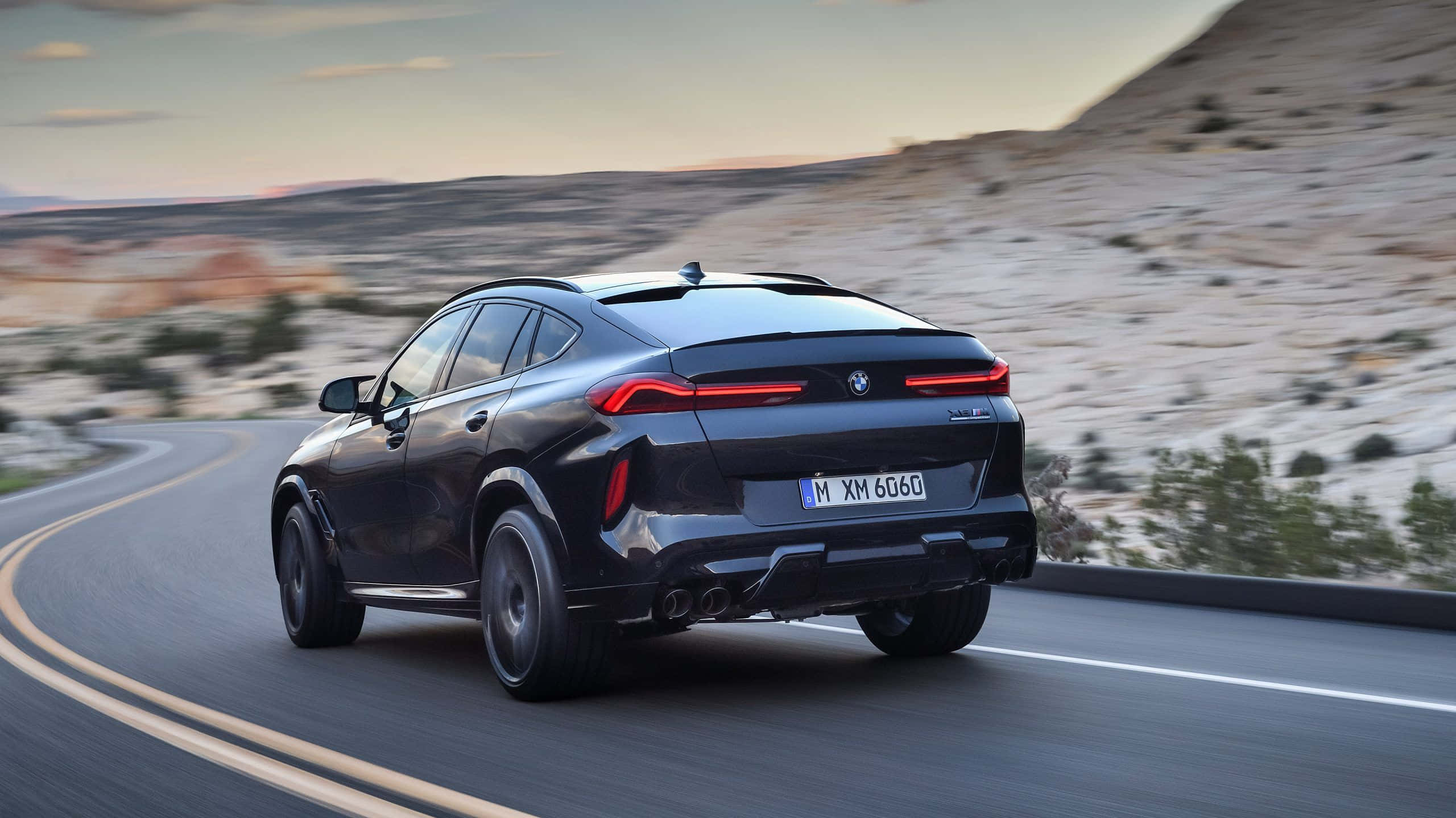 探索BMW X6 M，體驗刺激駕馭樂趣 壁紙 — 一輛時尚的黑色BMW XM SUV在日落時分沿著蜿蜒的沙漠公路行駛，帶有模糊的動態和溫暖的金色光線。