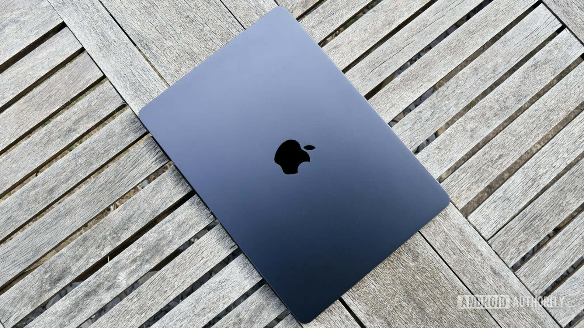 探索MacBook的無限可能 壁紙 — 一部時尚的深藍色 MacBook 合上放在經風化的灰色木板上，在自然日光下。