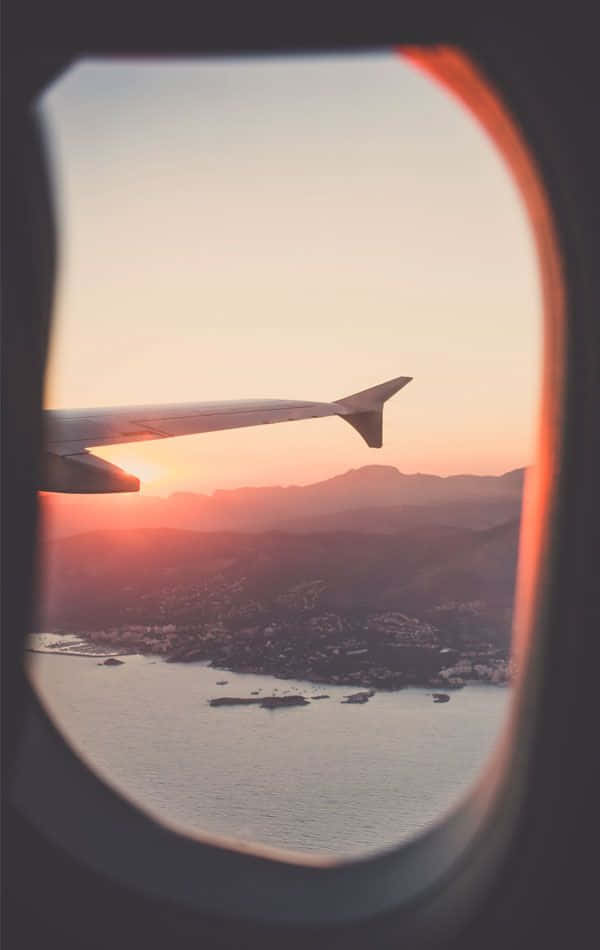 探索世界，擁抱不同文化 壁紙 — 透過圓形飛機窗戶看到的飛機機翼在溫暖的日落映襯下，剪影於沿海山脈和海洋之上。