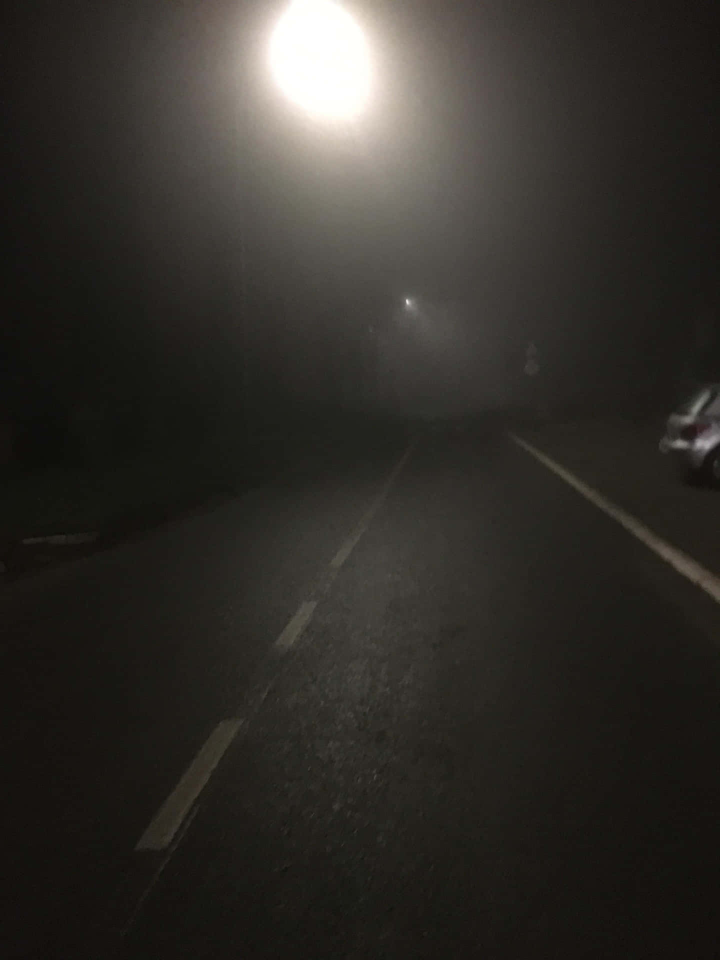 夜晚一條黑暗霧濛濛的道路，由一盞街燈照亮，旁邊可見一輛汽車。