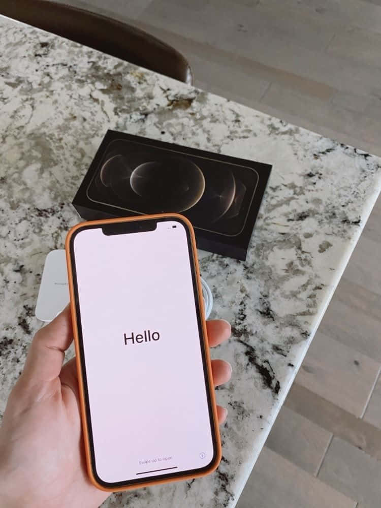 探索全新 iPhone 12 壁紙 — 一隻手喺雲石枱面上，用橙色機殼嘅新iPhone喺原裝盒上面顯示Hello畫面。
