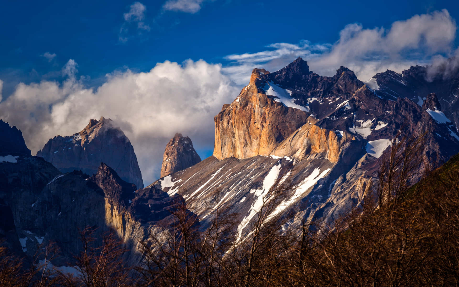 探索巴塔哥尼亞的壯麗景觀 壁紙 — 陽光照射的花崗岩山峰穿透戲劇性的雲層，覆蓋著雪的山脊，前景是光禿禿的樹木。
