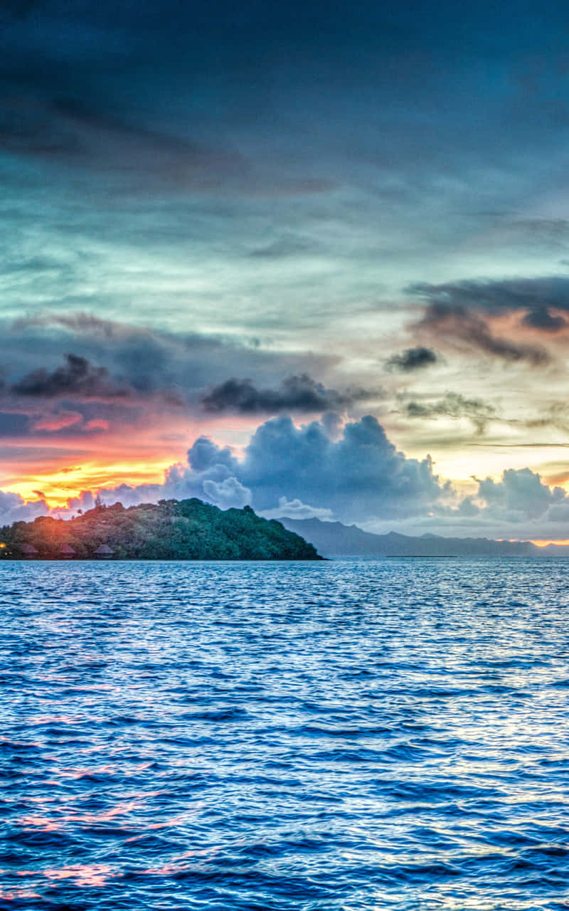 探索未受破壞的波拉波拉天堂 壁紙 — 夕陽西下，天空被染成火橙色和深藍色，平靜的海洋上浮現出一個鬱鬱蔥蔥的綠色島嶼輪廓。