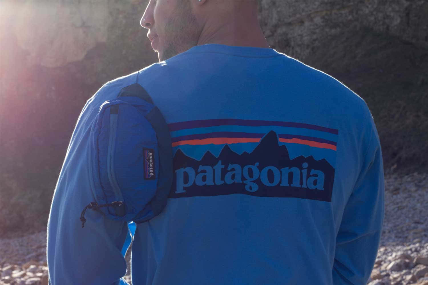 探索未經開發的巴塔哥尼亞之美 壁紙 — 一名身穿藍色Patagonia襯衫的男子，腰間掛著腰包，站在陽光普照的岩石背景前。