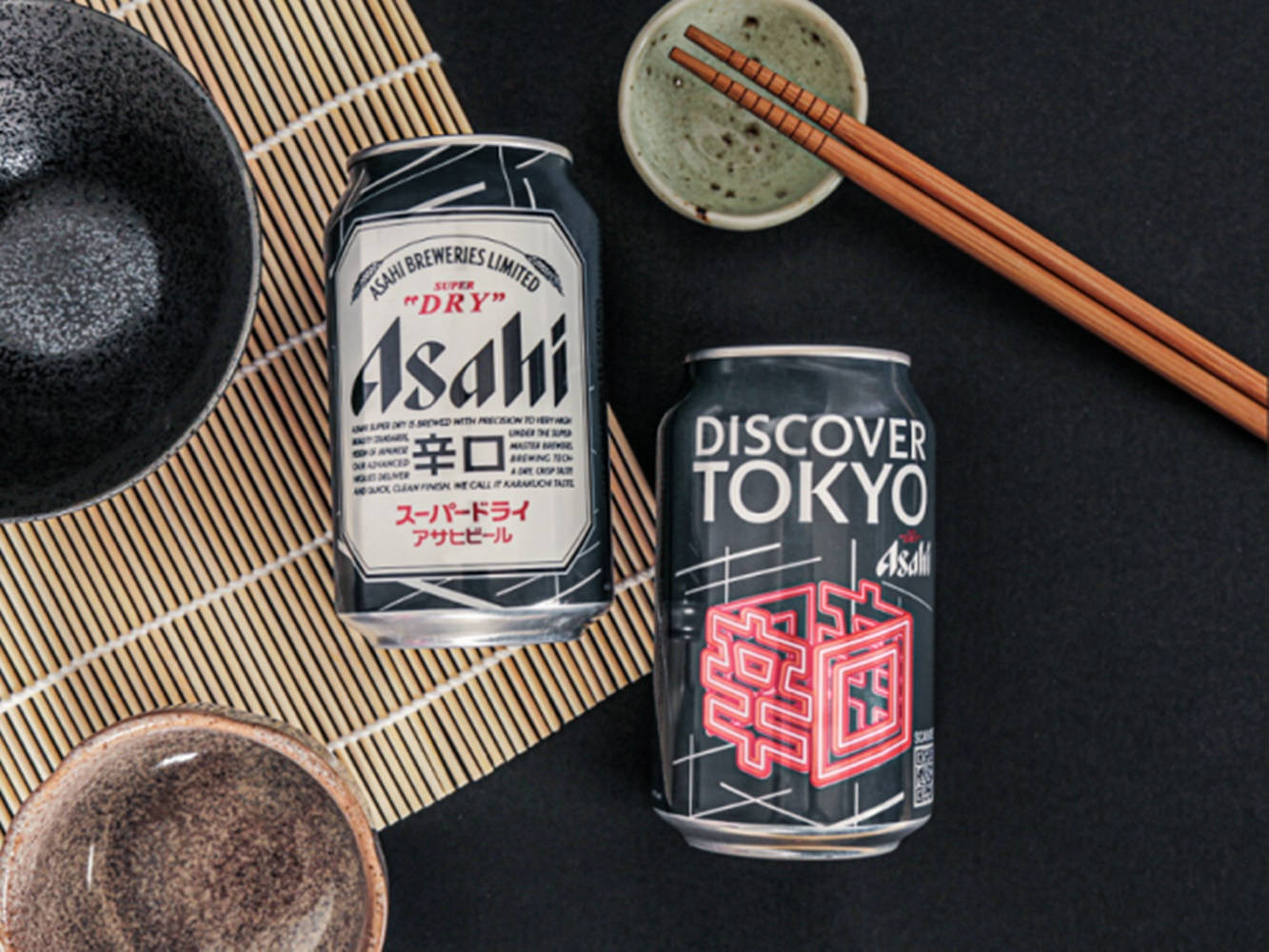 探索東京朝日超干限定版 壁紙 — 兩罐印有日文字樣的Asahi啤酒罐放在竹蓆上，旁邊有陶瓷碗和木筷子。