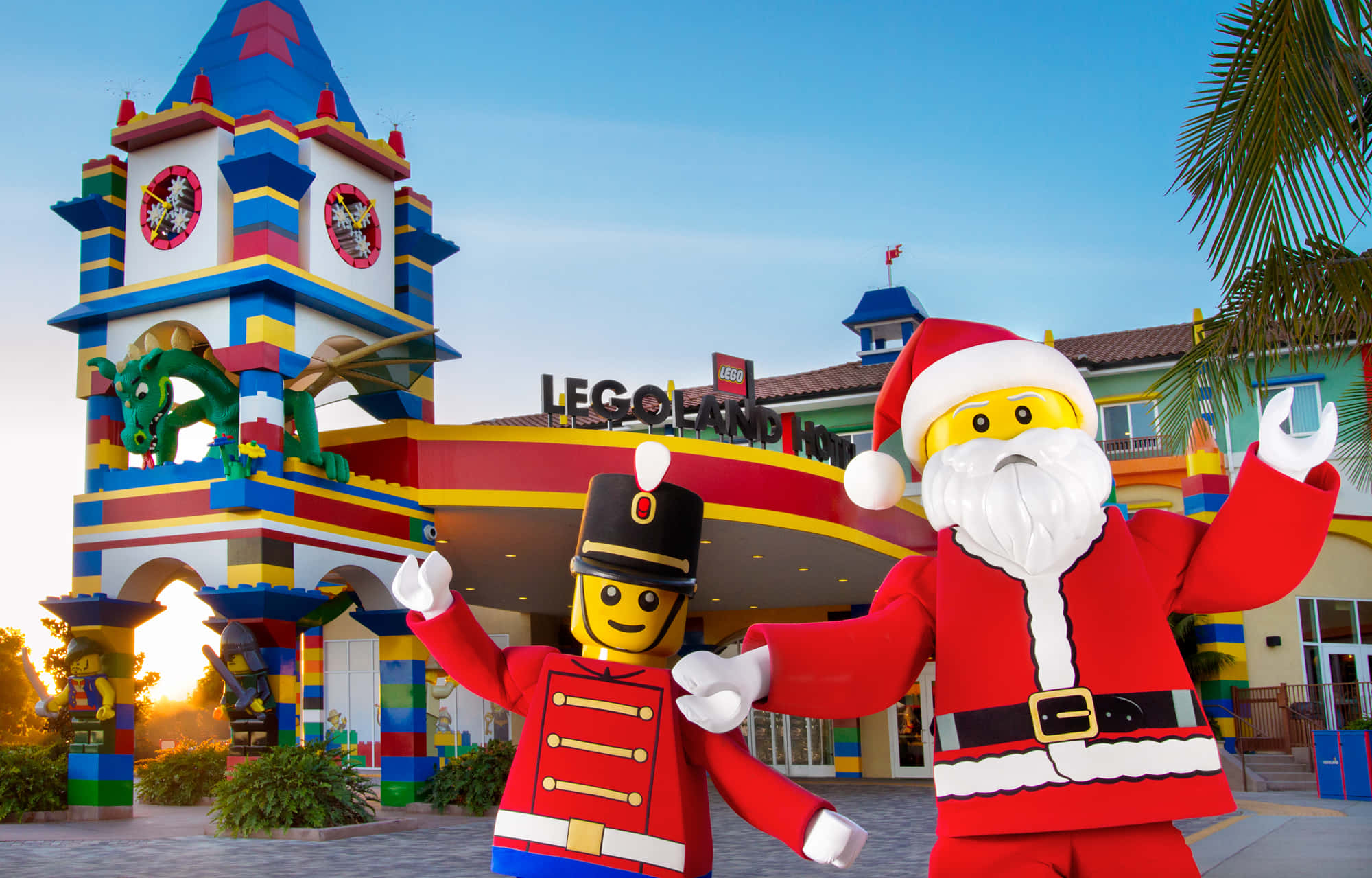 探索樂高樂園的神奇世界！ 壁紙 — 兩個巨型LEGO公仔，一個士兵和聖誕老人，在日落時分愉快地站在色彩繽紛的LEGO主題遊樂園入口前。