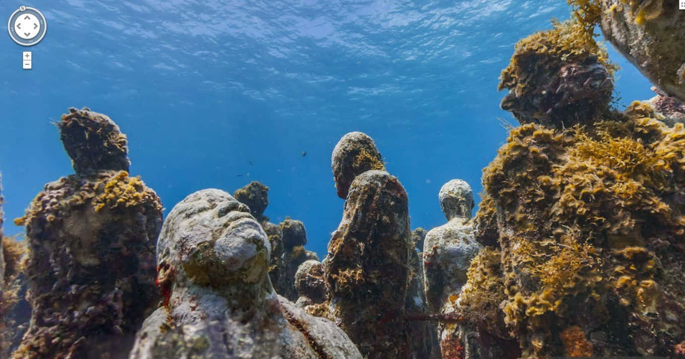 探索水下恐懼海洋的神秘深處 壁紙 — 被珊瑚和海藻覆蓋的水底雕像從清澈藍色海水下的海床升起。